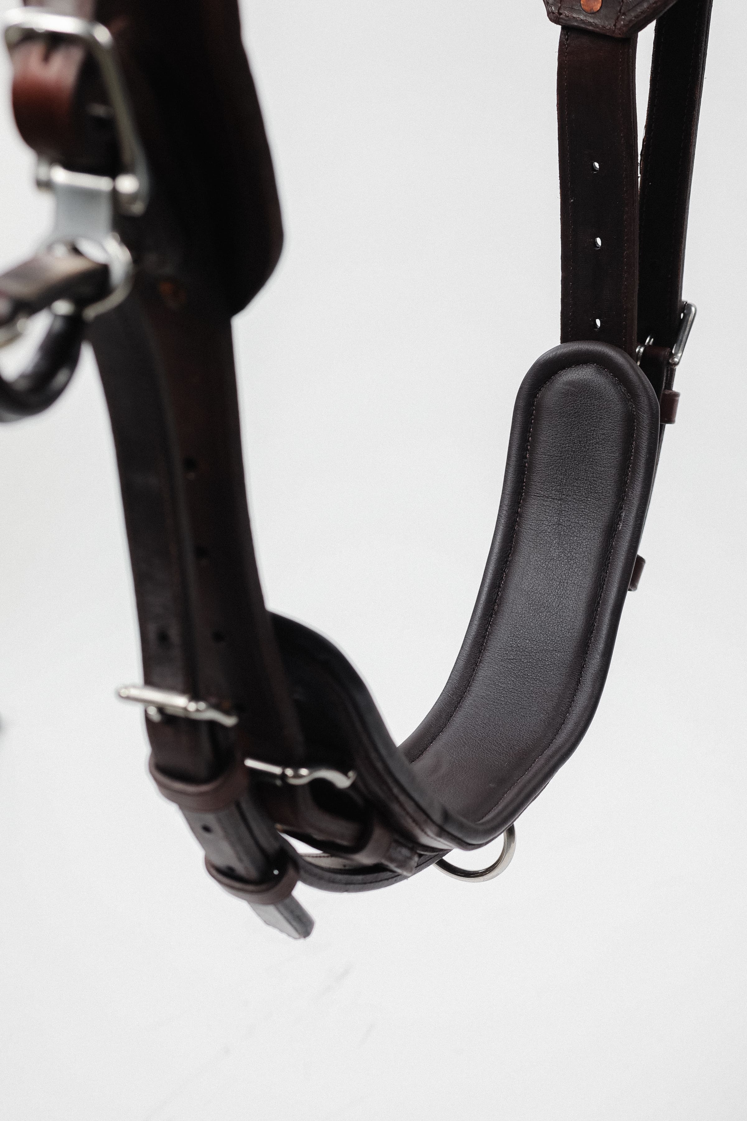 Close-up of a padded brown leather harness component, showing the texture of the leather, the stitching detail, and the metal buckles and rings, isolated on a white background.