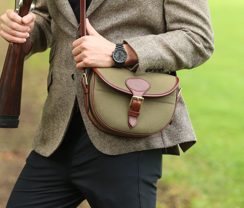 Man wearing a brown tweed jacket and holding a shotgun, carrying a Military Green Ashcombe Cartridge Bag with Havana brown leather trim, made by Freedman’s Harness.