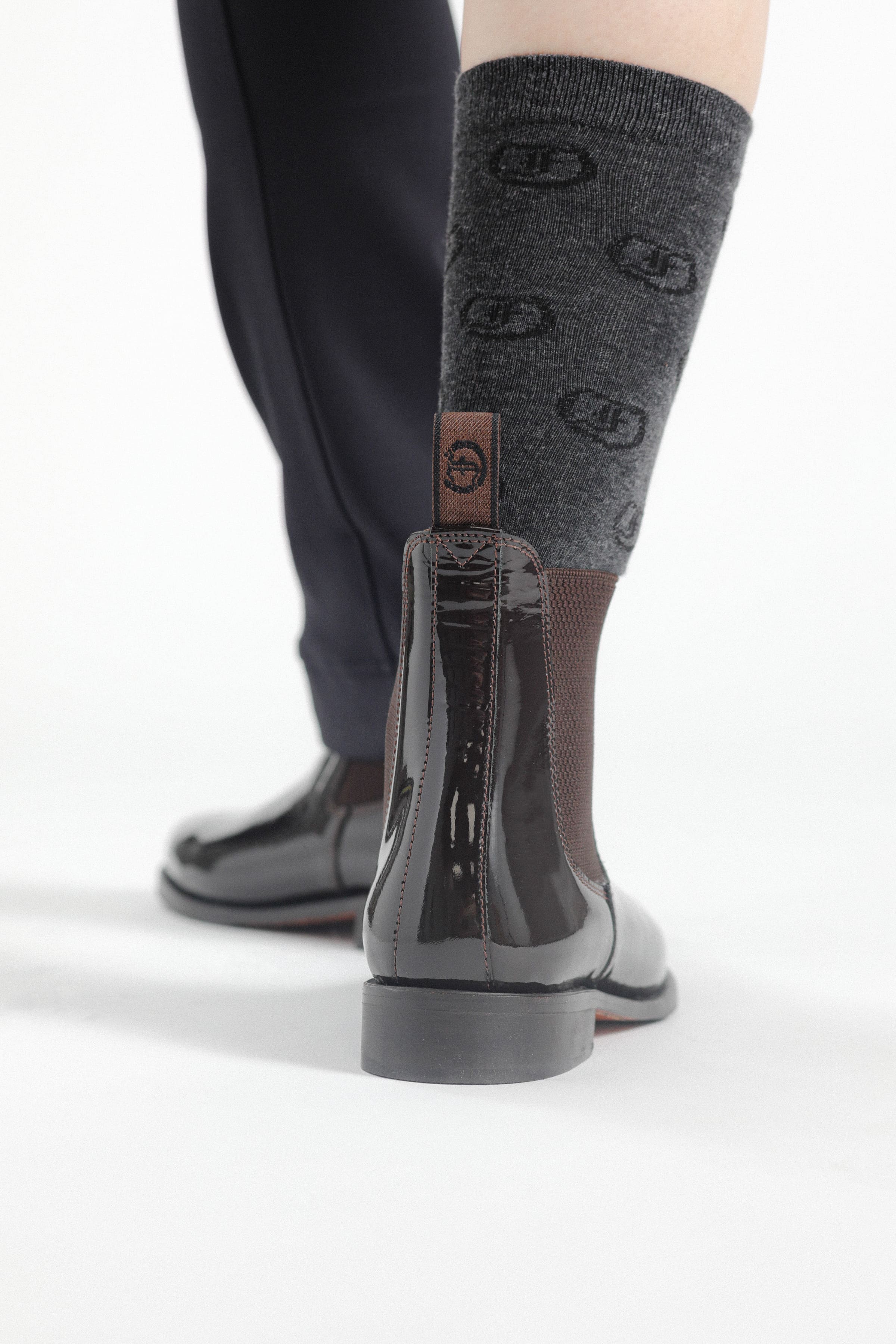 Single black patent leather Adelaide Show Jodhpur Boot on a person's foot, with patterned socks visible, from a rear perspective.