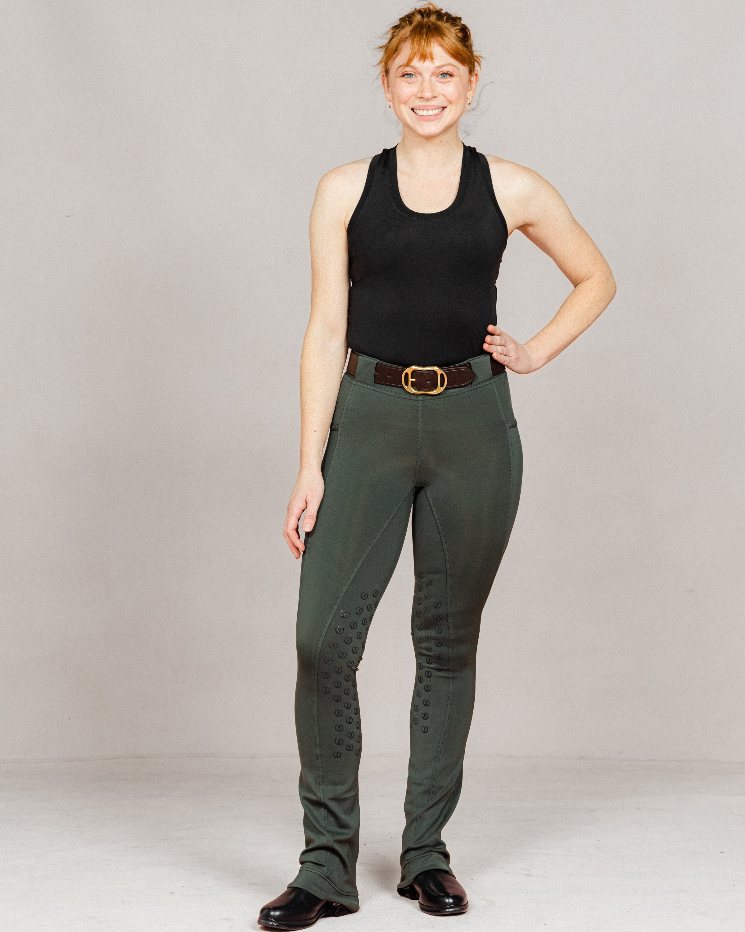 Woman wearing Umber Green jodhpurs by Freedman Harness with full seat grip, brown leather belt, and black riding boots – stylish equestrian outfit for everyday riding