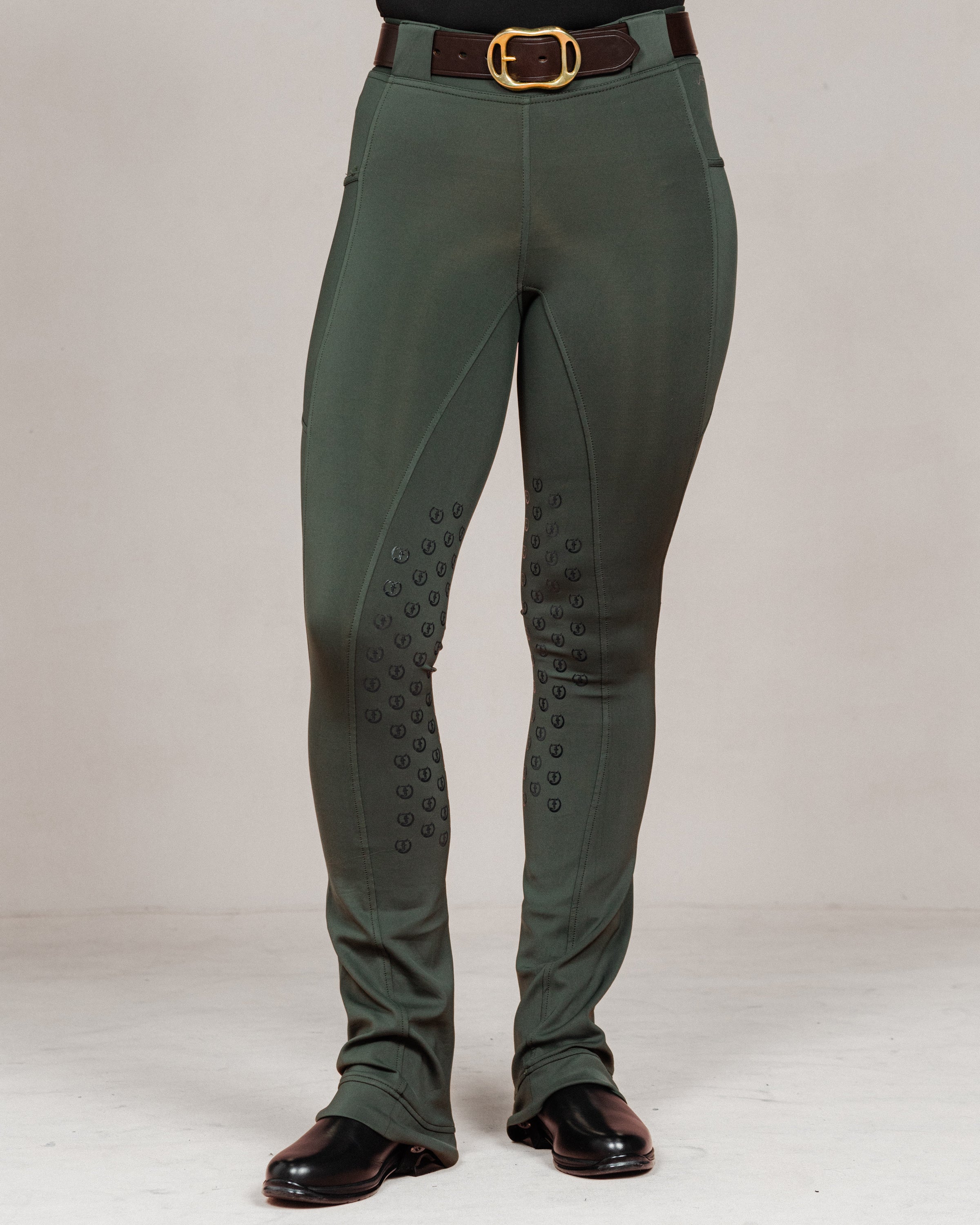 Front view of women's Umber Green jodhpurs by Freedman Harness, featuring full seat silicone grip, flared leg, brown belt with gold buckle, and black riding boots – high-performance equestrian pant