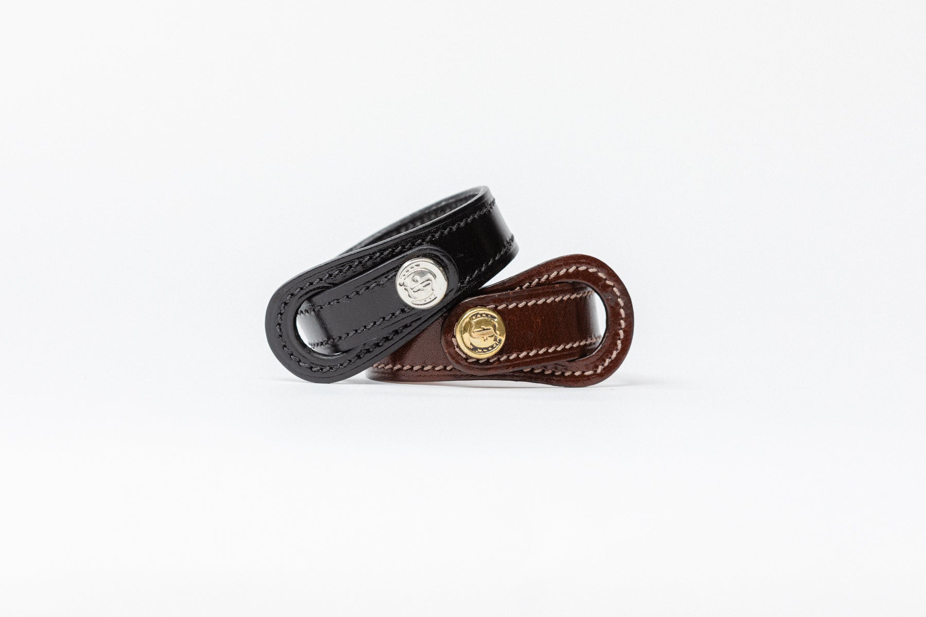 Pair of Signature Leather Bracelets by Freedman's, one black with silver logo button and one brown with brass logo button, both with white stitching, displayed on a white background.