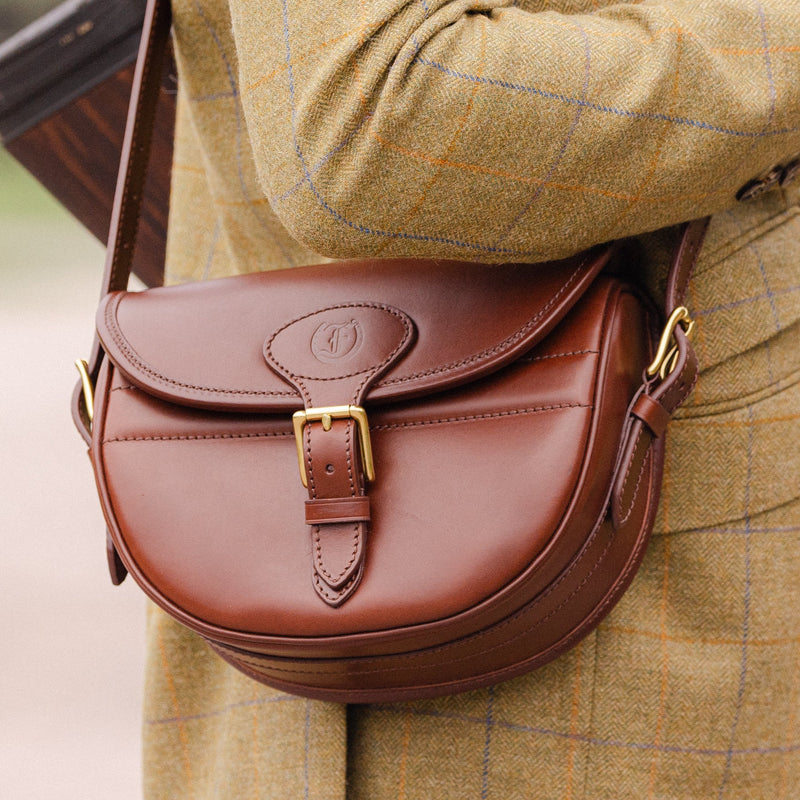 Close-up of the Ashcombe Cartridge Bag in Havana brown leather with brass buckle hardware, worn over a green tweed jacket, made by Freedman’s Harness.
