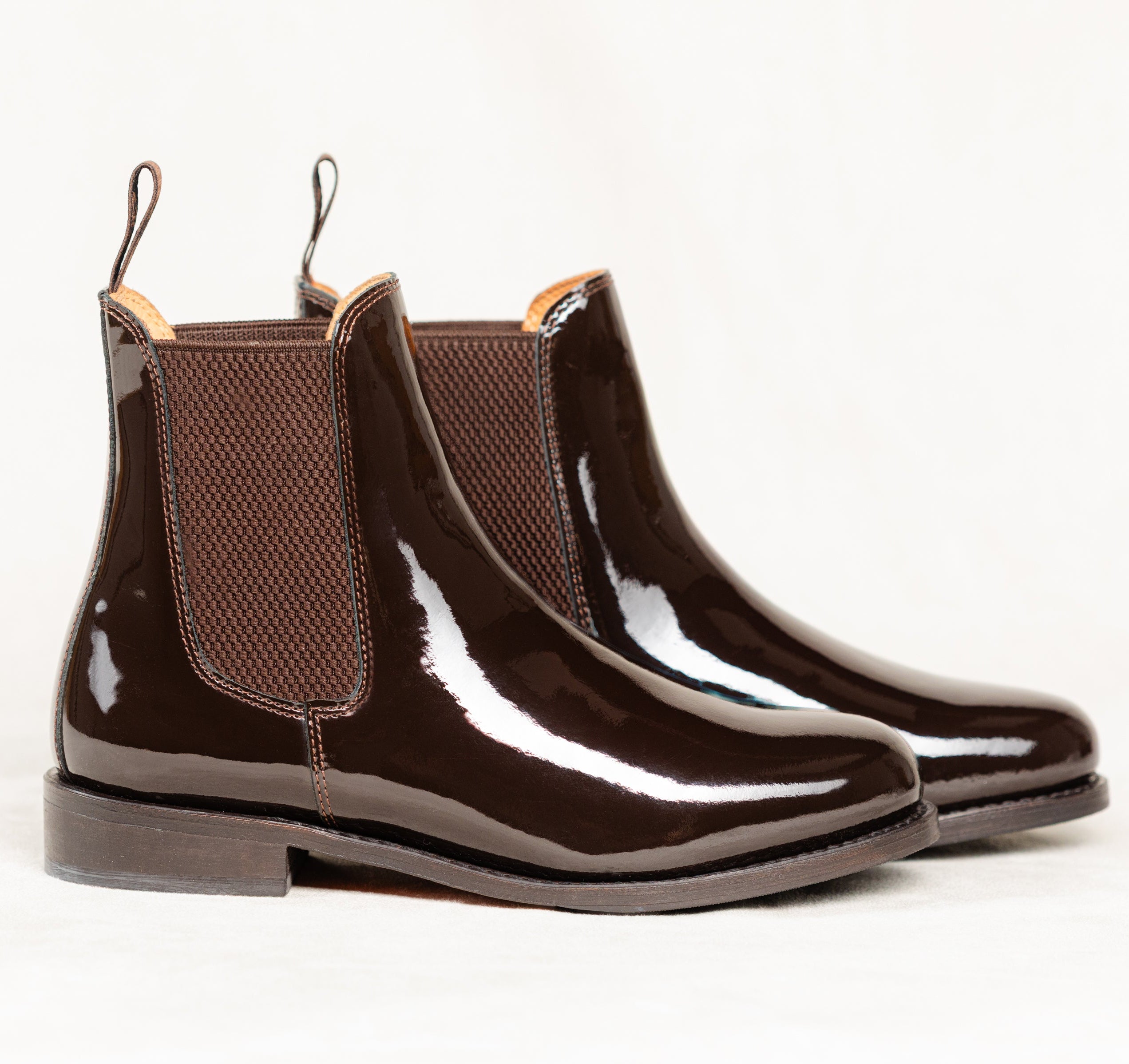 Pair of Adelaide Patent Show Jodhpur Boots, designed for equestrian showing, made of Brown patent leather with elastic ankle panels and low heels, isolated on a white background.