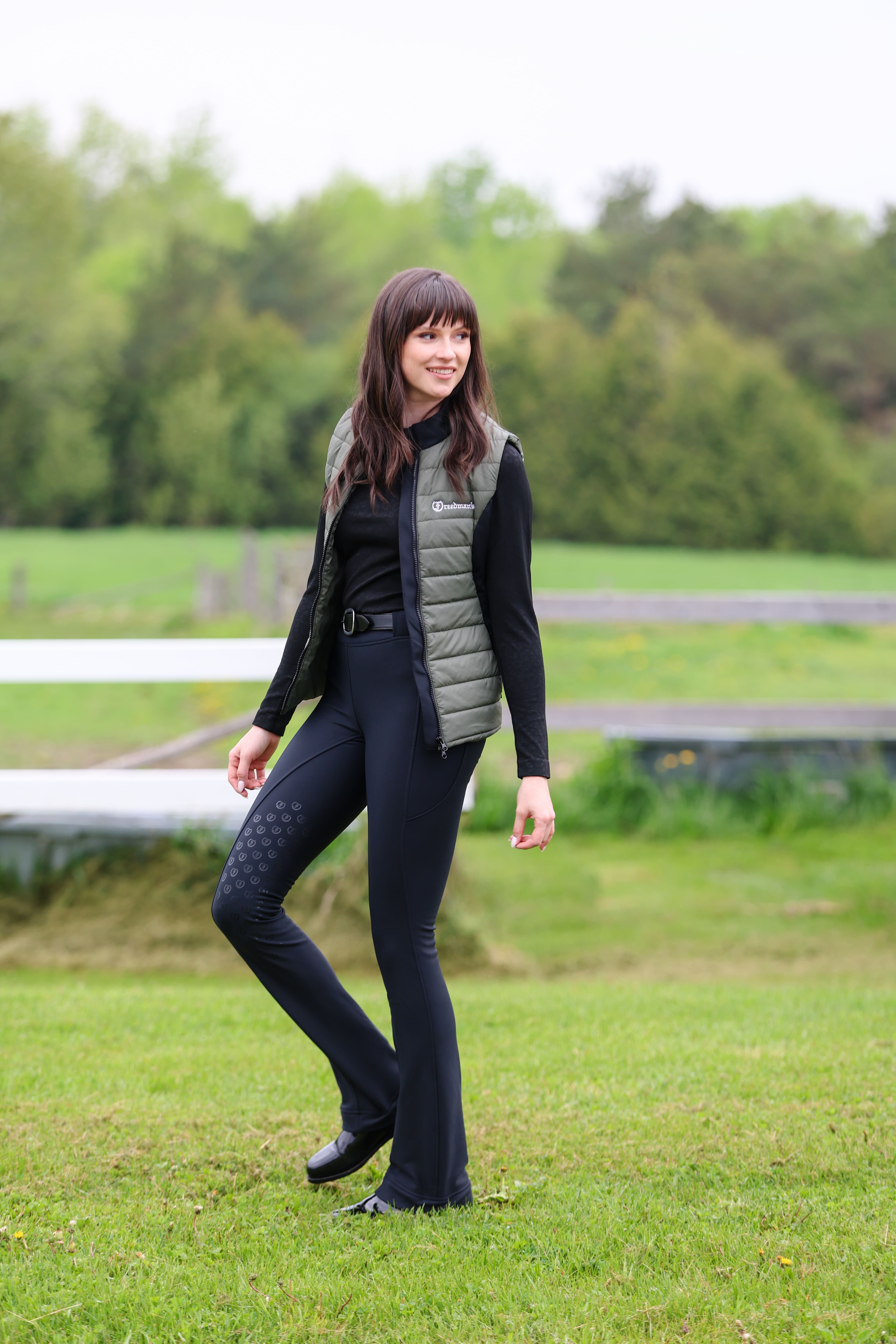 Ladies Essential Quilted Vest by Freedman's in Military Green with Black side accent and black zipper detail, modeled by smiling woman with dark long hair wearing slate black top and black jods against natural background, showing athletic apparel fit and style