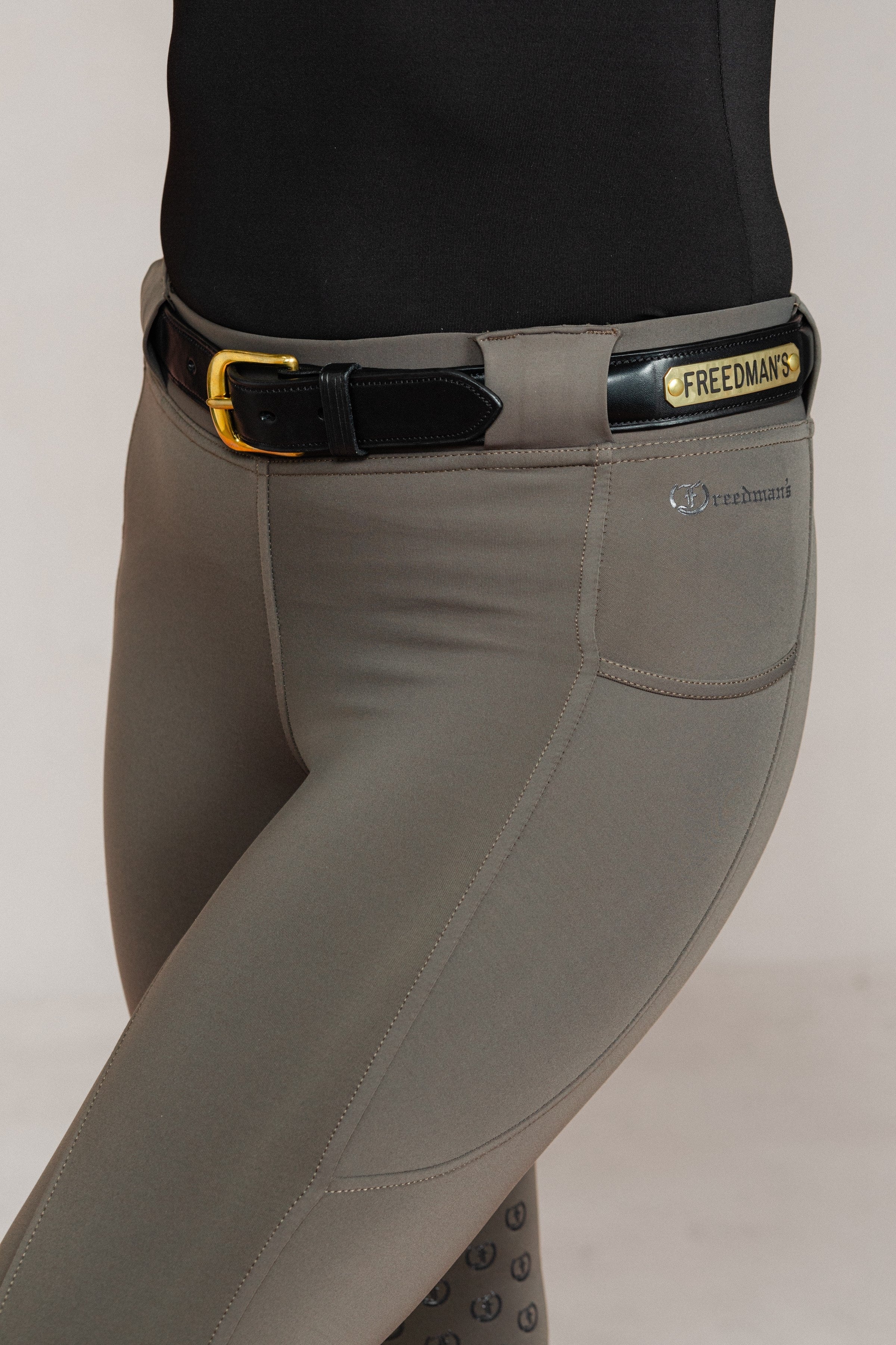Raised Custom Name Plate Belt in black leather with gold buckle and "FREEDMAN'S" nameplate, worn with grey equestrian riding pants.