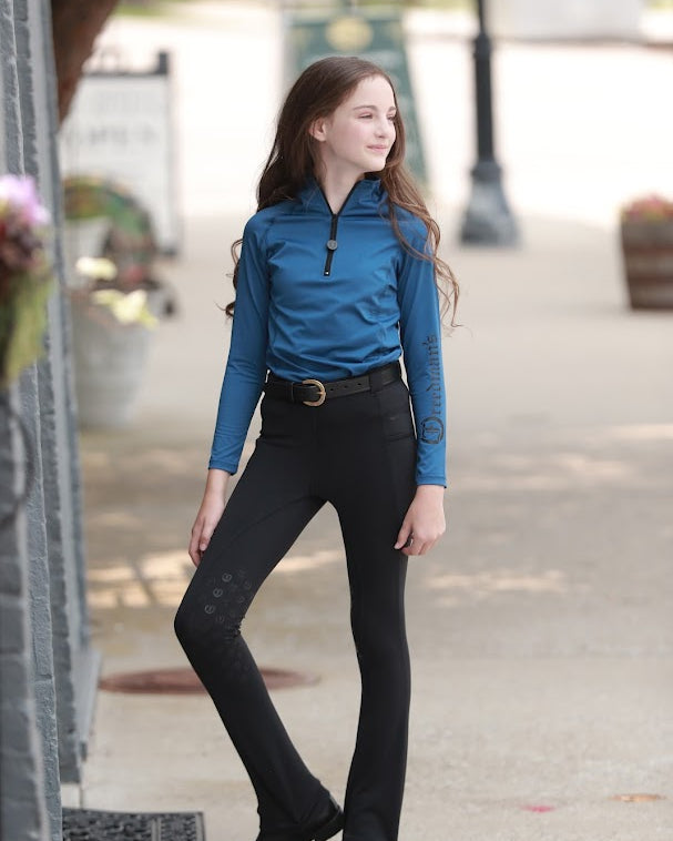 Young girl wearing the Freedman's Kids 1/4 Zip Base Layer in blue, paired with black riding breeches, a belt, and black boots. She stands confidently outdoors, showcasing the stylish and functional equestrian top with a sleek zip-up design and Freedman’s logo on the sleeve. Perfect for riding and active wear.