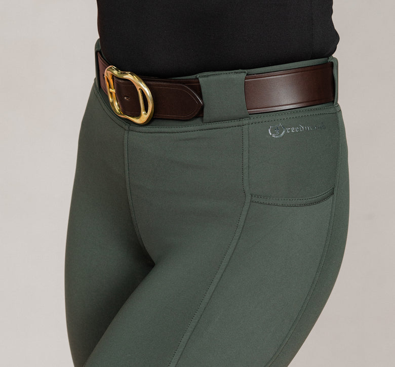 Detail view of women's Umber Green jodhpurs by Freedman Harness, featuring front pocket, embroidered logo, and brown leather belt with gold buckle