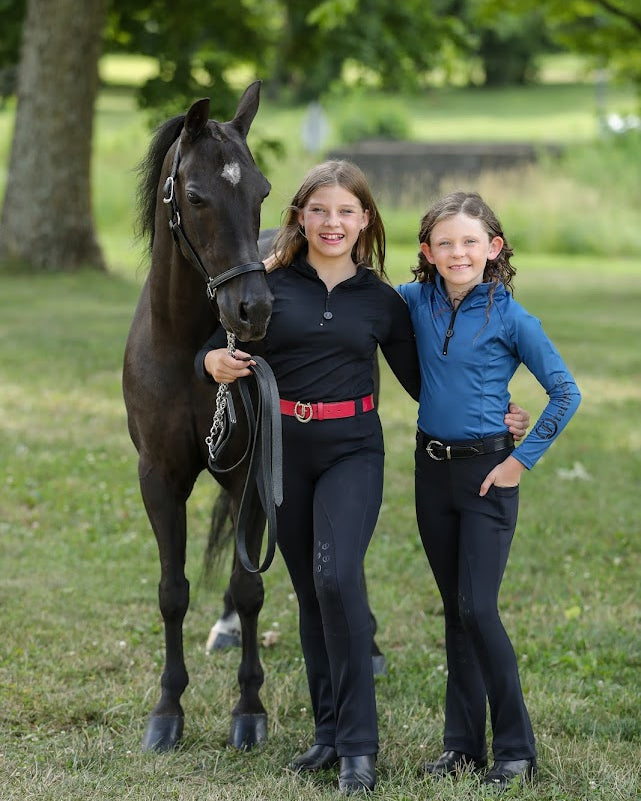 Two Young girls wearing the Freedman's Kids 1/4 Zip Base Layer one in blue and other in black, paired with black riding breeches, a belt, and black boots. She stands confidently outdoors, showcasing the stylish and functional equestrian top with a sleek zip-up design and Freedman’s logo on the sleeve. Perfect for riding and active wear.