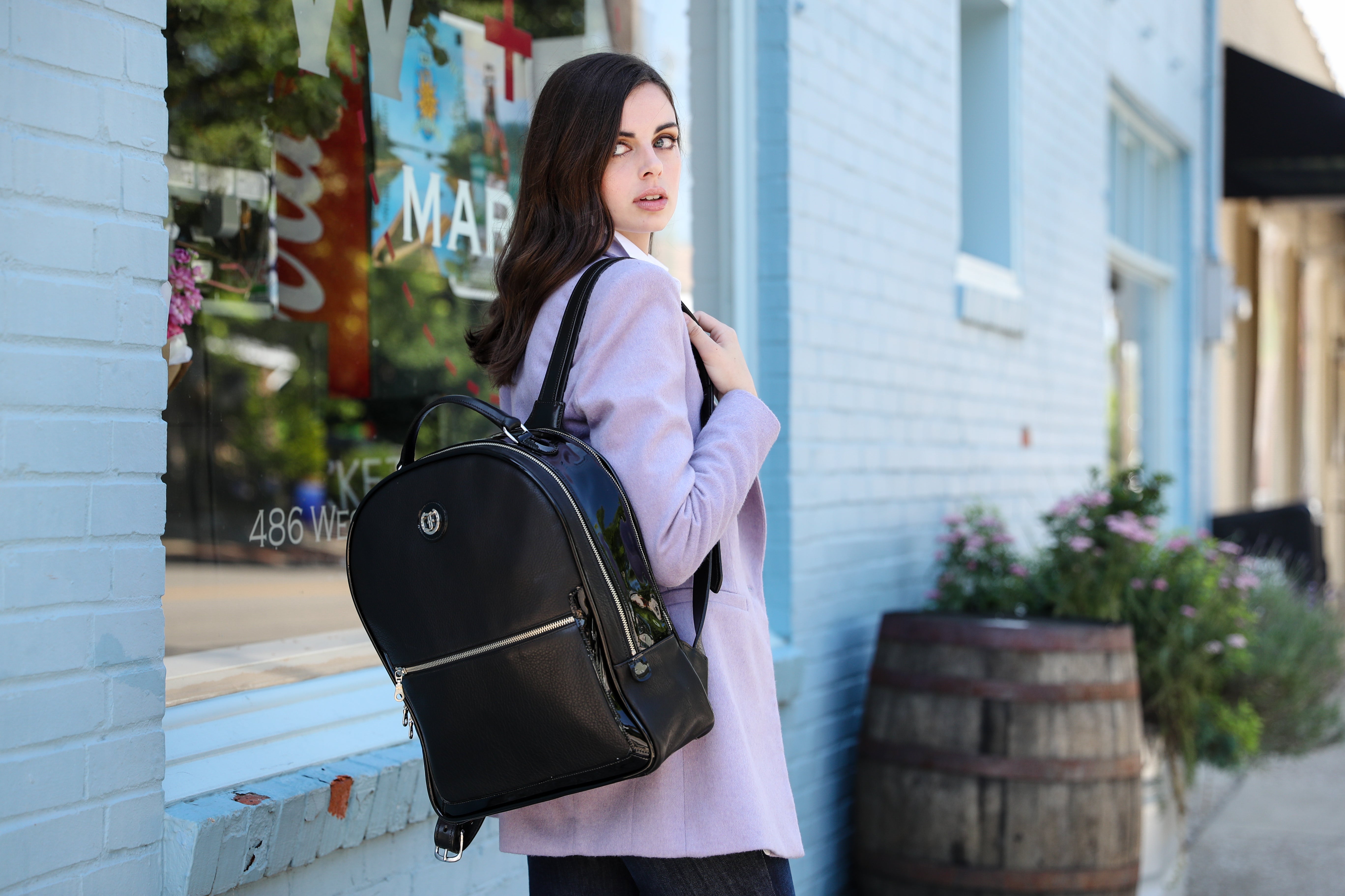 Stylish woman wearing a lilac blazer and carrying 'The Jaunty Backpack' by Freedman, a sleek black leather backpack with silver accents, walking past a charming blue storefront with flowers and a rustic barrel in the background