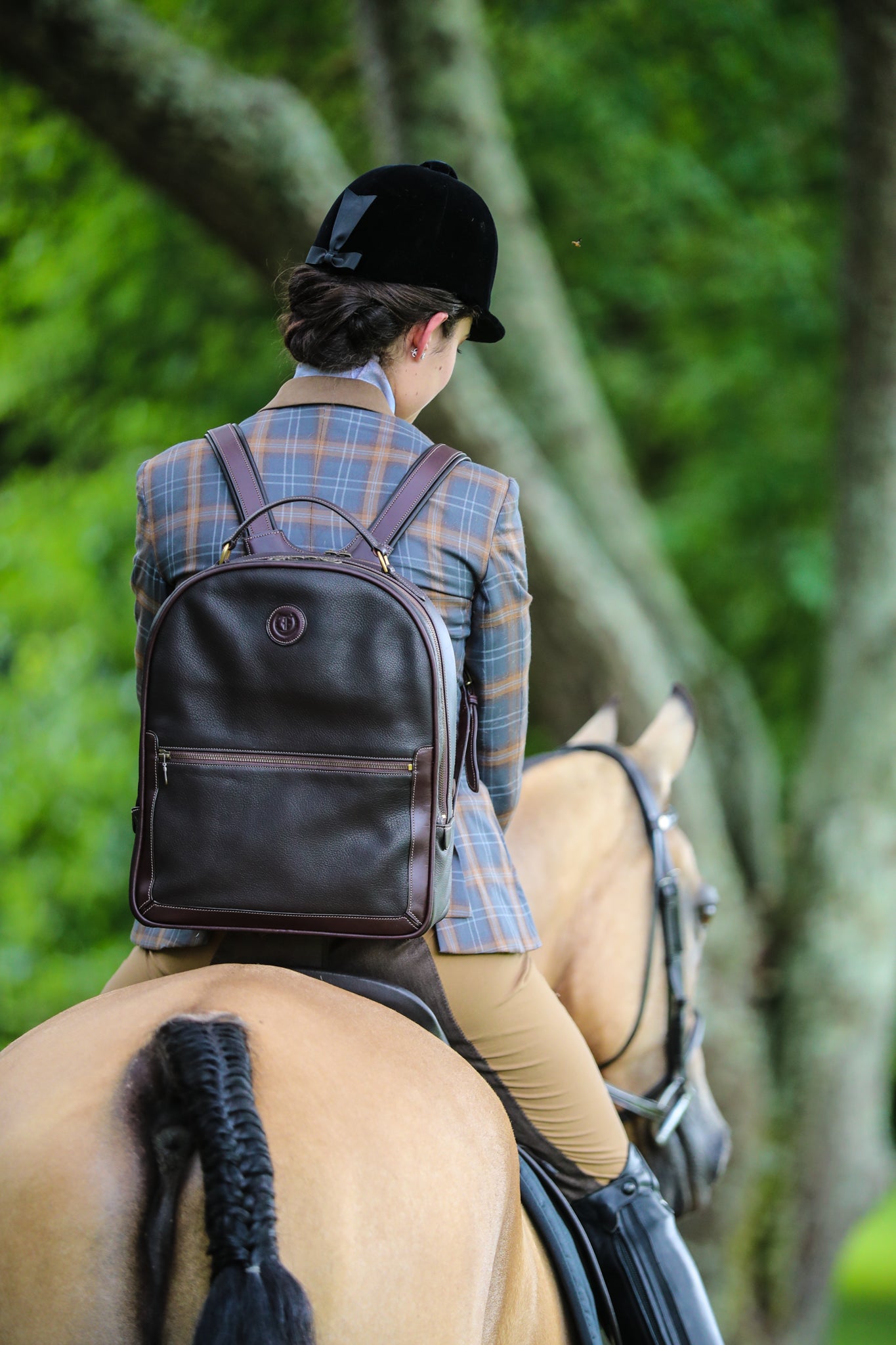 Equestrian rider wearing a plaid jacket and black velvet helmet, carrying 'The Jaunty Backpack' by Freedman—a sophisticated brown leather backpack with gold accents—while riding a beautifully groomed horse through a lush green setting