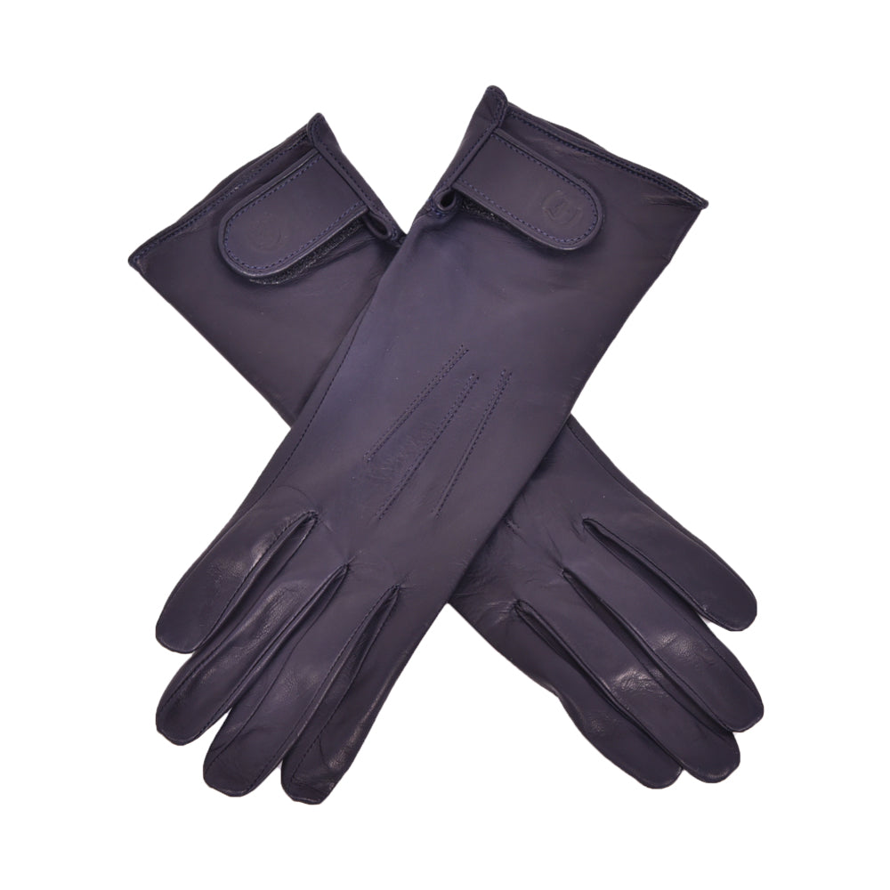 The Yorkville Show Glove, dark navy leather gloves crossed, with wrist strap and button detail, visible seam stitching, light background.