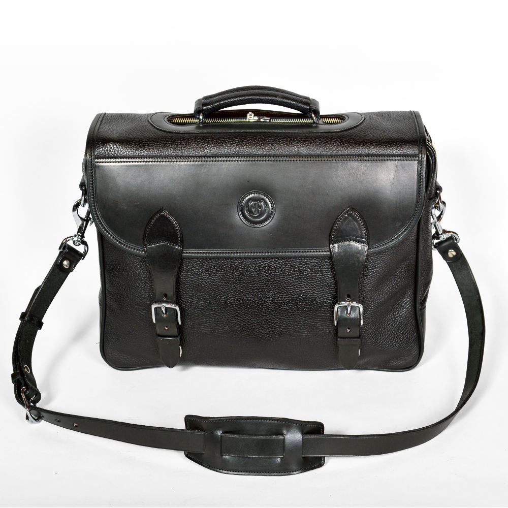 Wagonette Briefcase, black textured leather, flap closure with dual buckle straps, silver hardware, embossed circular logo on front, detachable shoulder strap with padded section, visible zipper closure at top, white background.