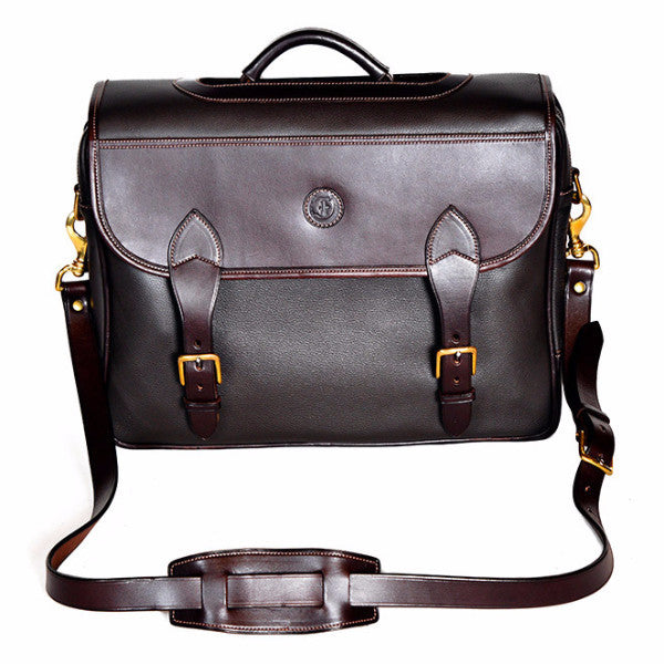 Wagonette Briefcase, dark brown textured leather, flap closure with dual buckle straps, silver hardware, contrast stitching, embossed circular logo on front, detachable shoulder strap with padded section, white background.