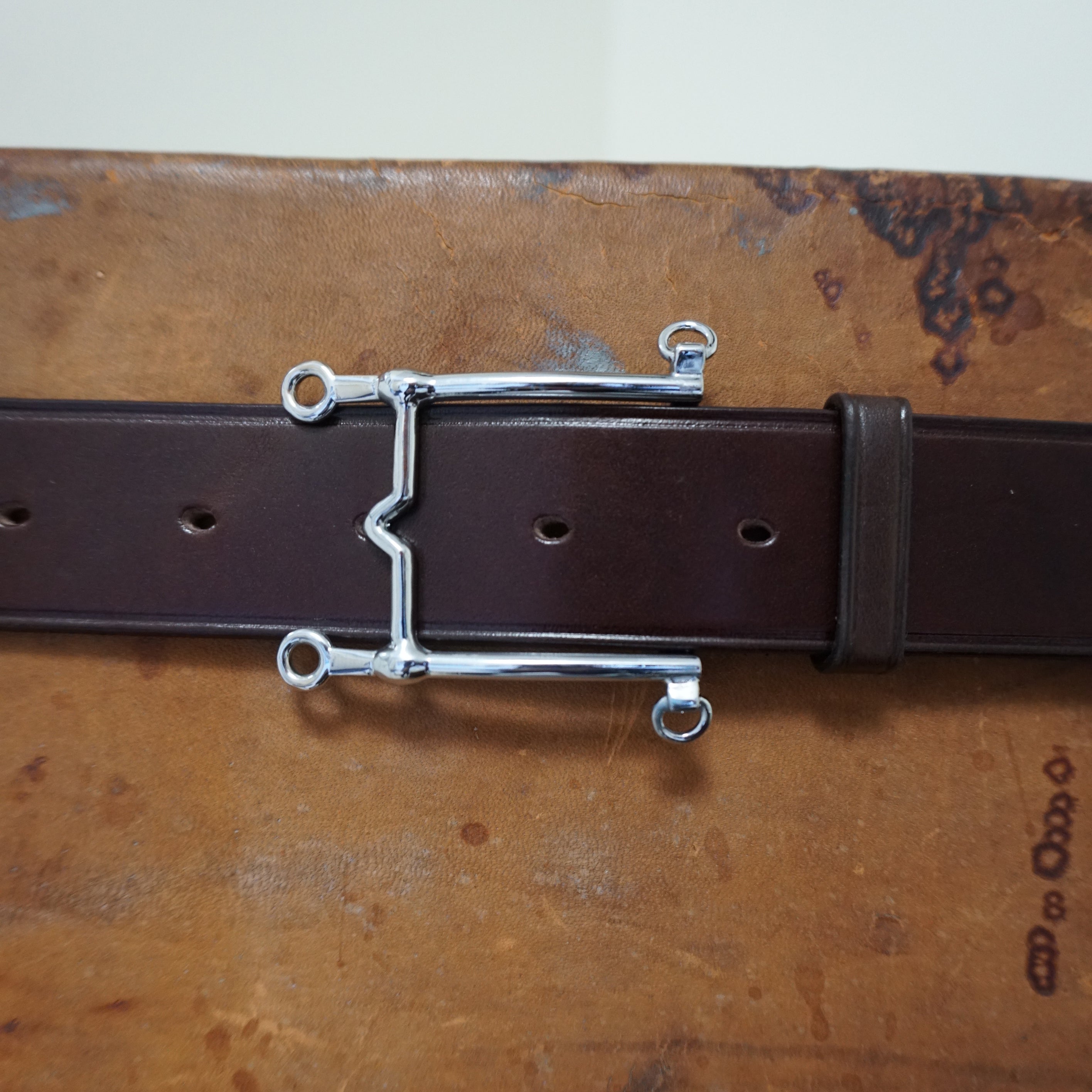 Silver bit buckle detail on brown leather belt, Weymouth model, shown on textured surface.