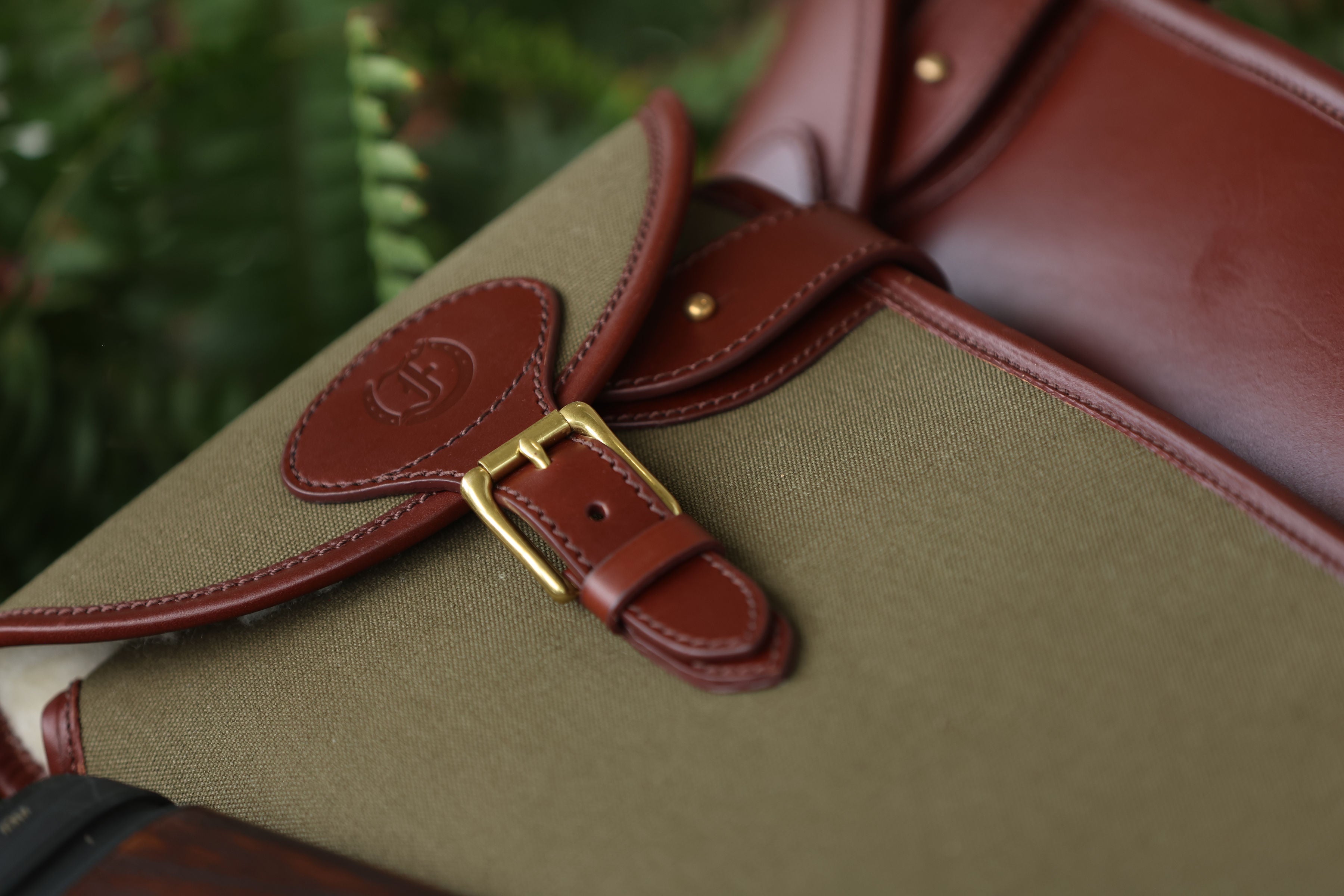 Close-up of a Sandringham Gun Slip strap and buckle with the Freedman’s Harness “F” logo, blurred green background.