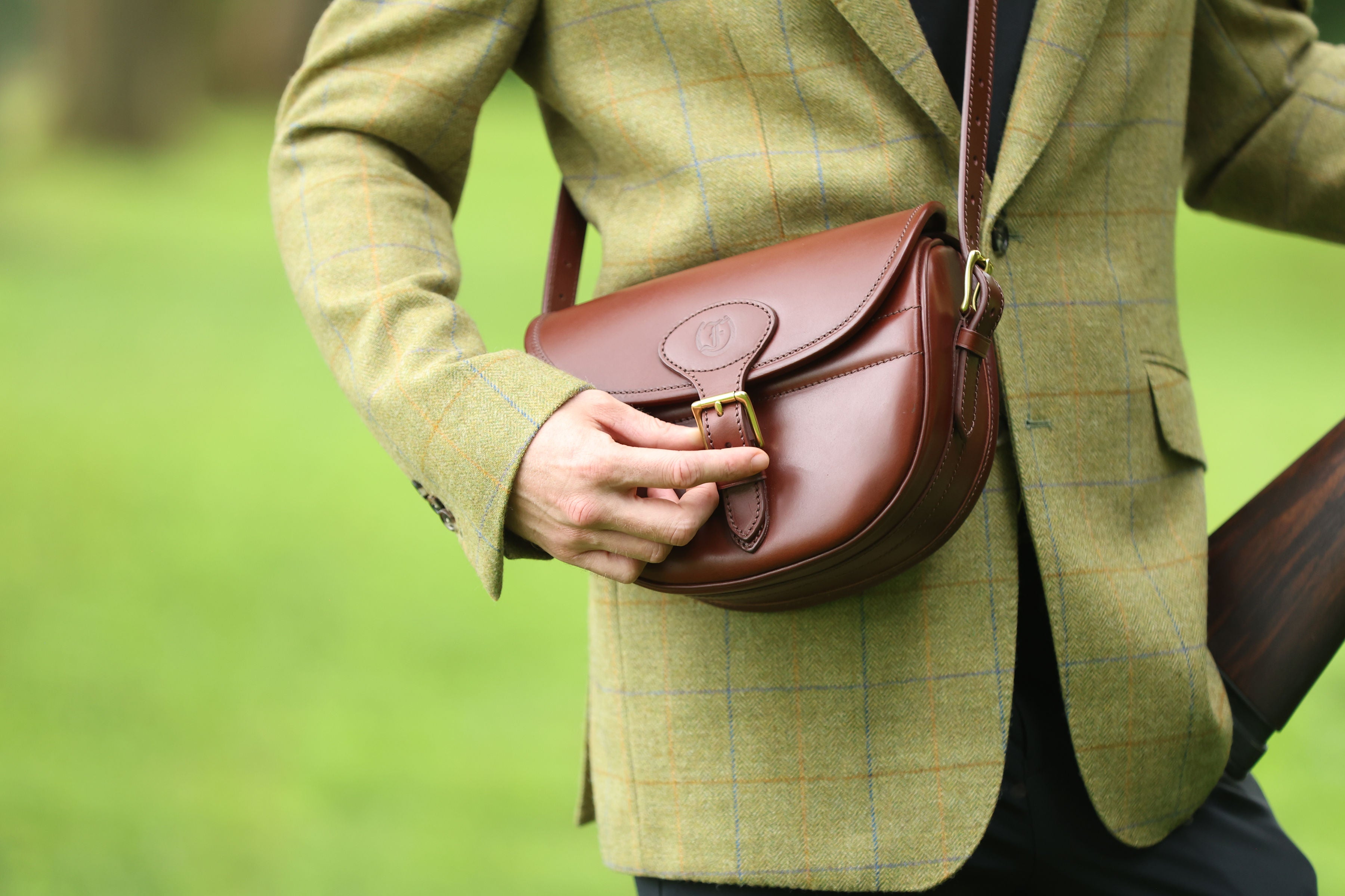 Man wearing a green tweed jacket adjusting the buckle on a Havana Brown Leather Ashcombe Cartridge Bag, made by Freedman’s Harness, in an outdoor setting.