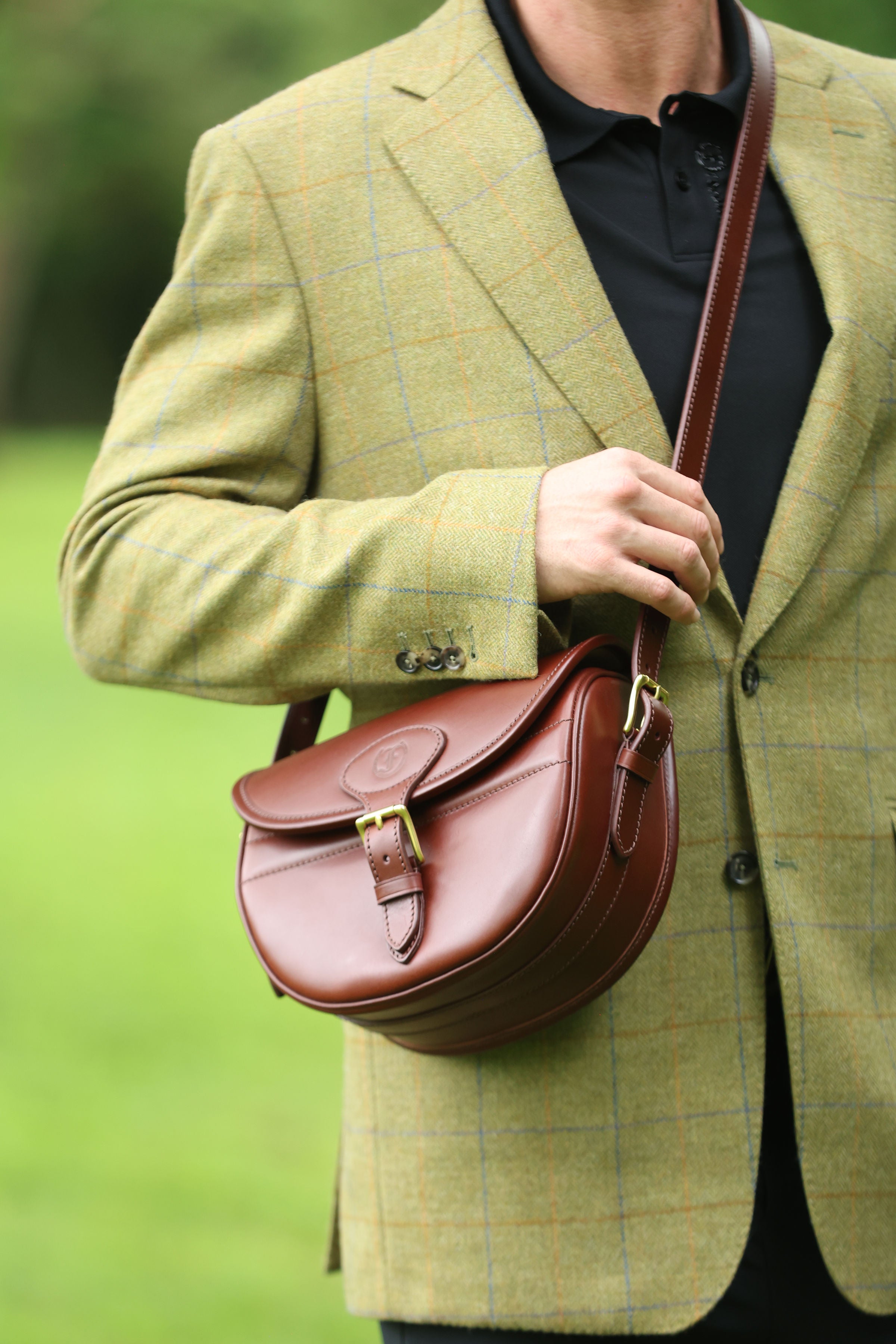 Man wearing a green tweed jacket adjusting the buckle on a Havana Brown Leather Ashcombe Cartridge Bag, made by Freedman’s Harness, in an outdoor setting.
