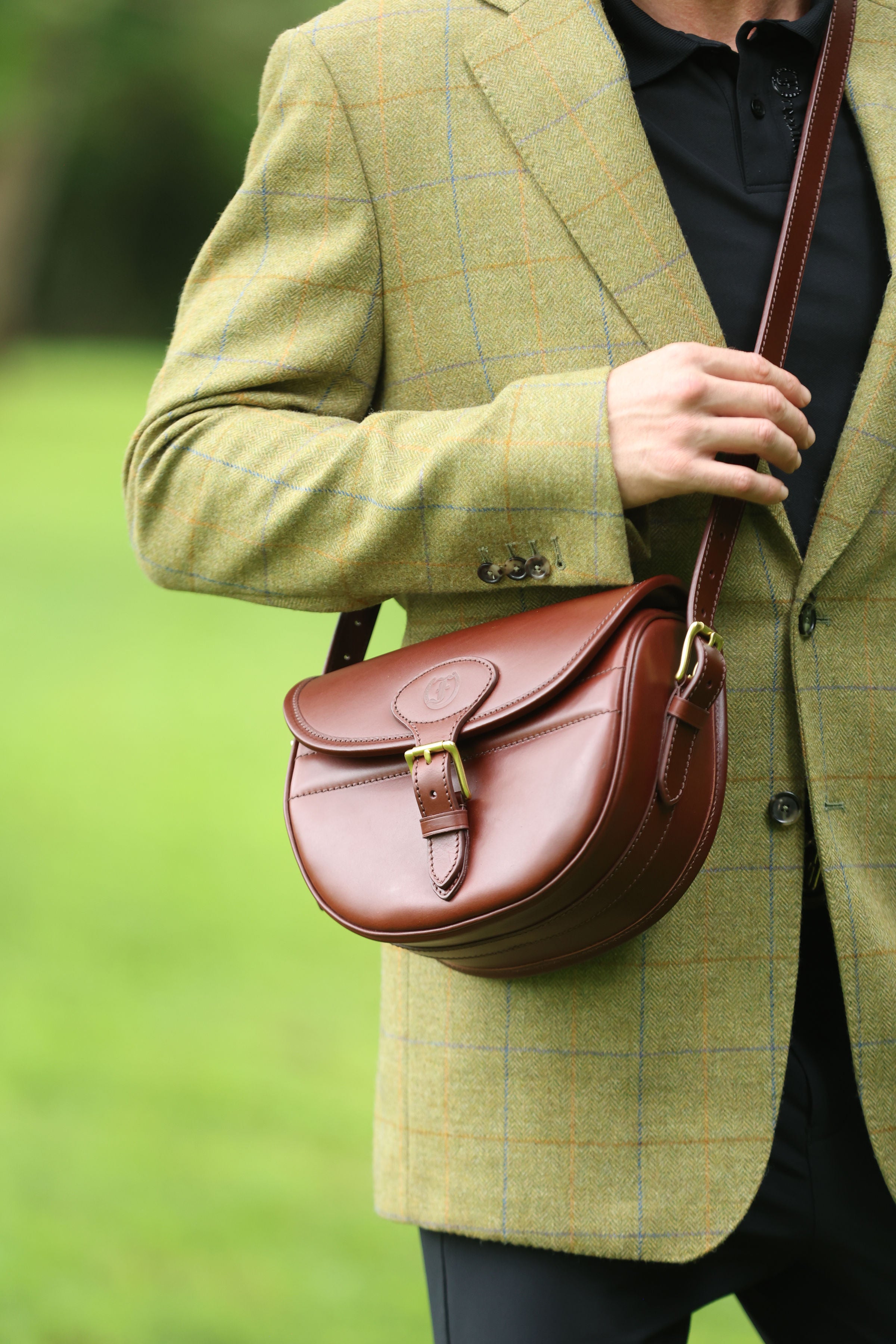 Man wearing a green tweed jacket adjusting the buckle on a Havana Brown Leather Ashcombe Cartridge Bag, made by Freedman’s Harness, in an outdoor setting.