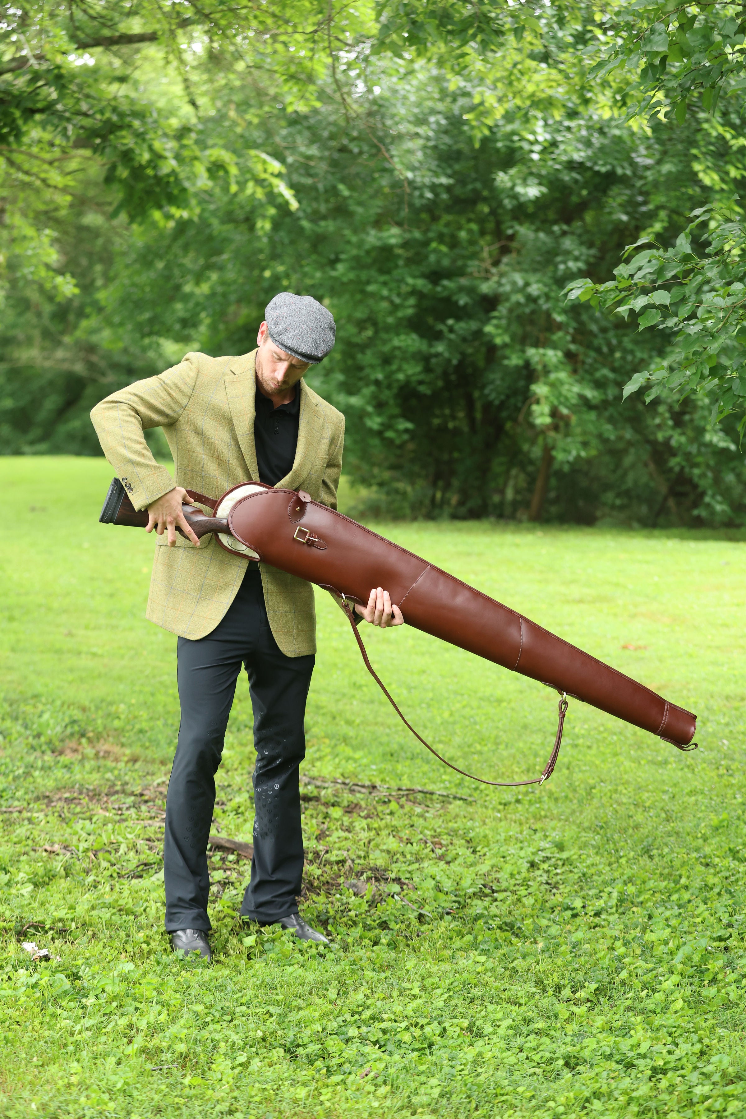 Man wearing a green tweed jacket and cap holding a Havana Brown Leather Sandringham Gun Slip outdoors on a grassy field, made by Freedman’s Harness.