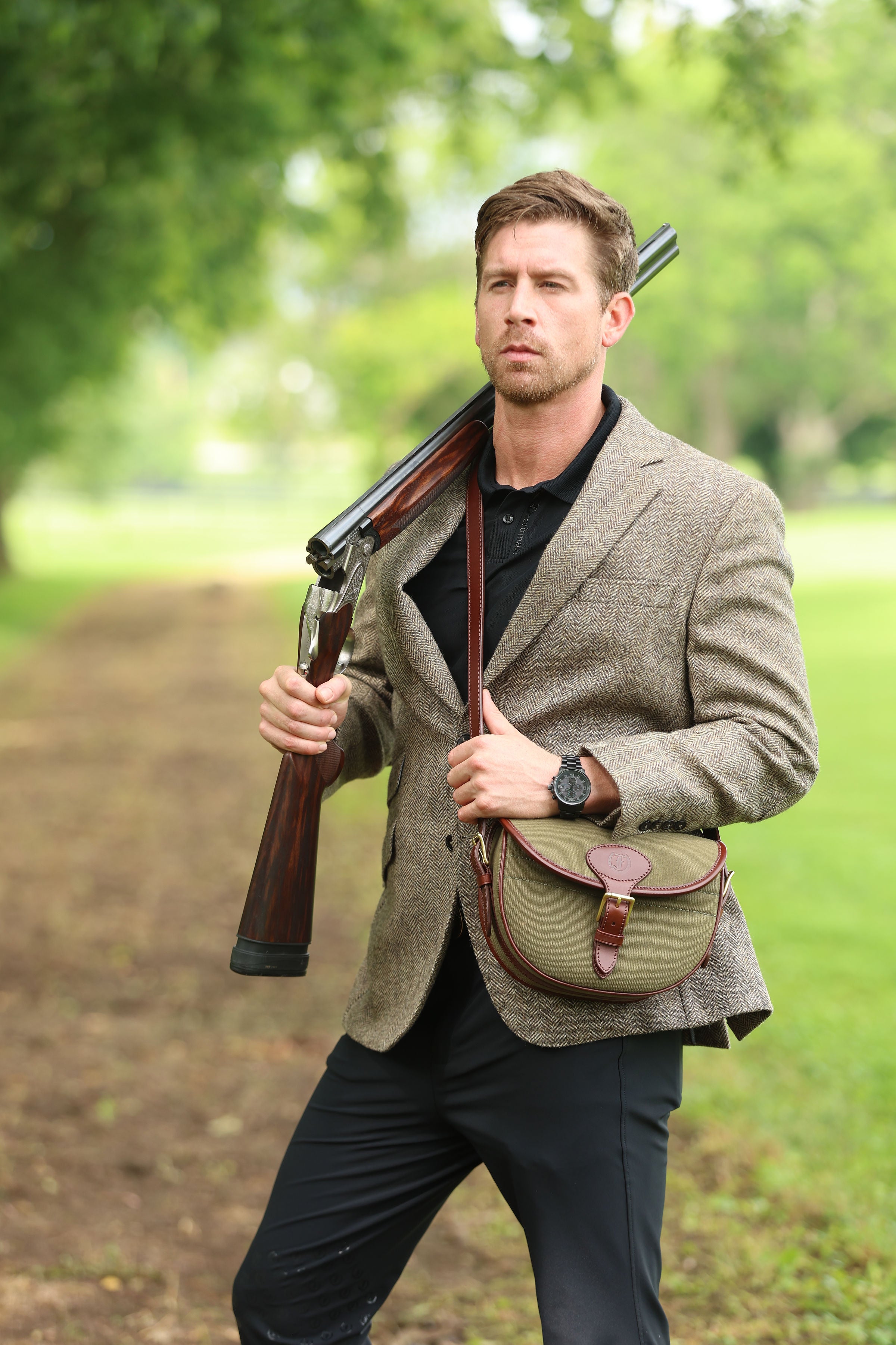 Man wearing a brown tweed jacket and black shirt carrying a shotgun over his shoulder with a Military Green Ashcombe Cartridge Bag featuring Havana brown leather trim, made by Freedman’s Harness.