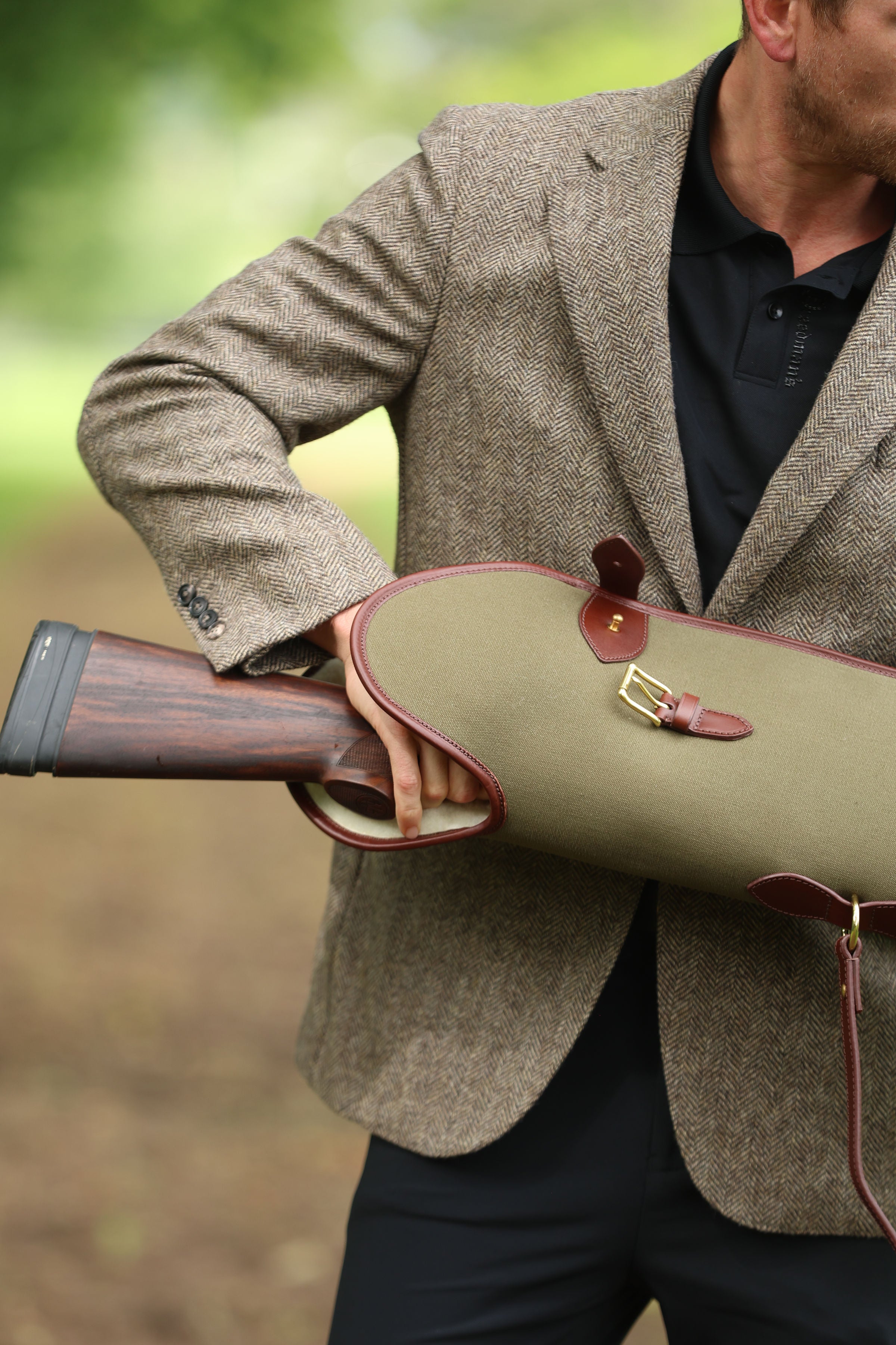Man in a brown tweed jacket holding a Military Green Sandringham Gun Slip with Havana brown leather trim in an outdoor setting, made by Freedman’s Harness.