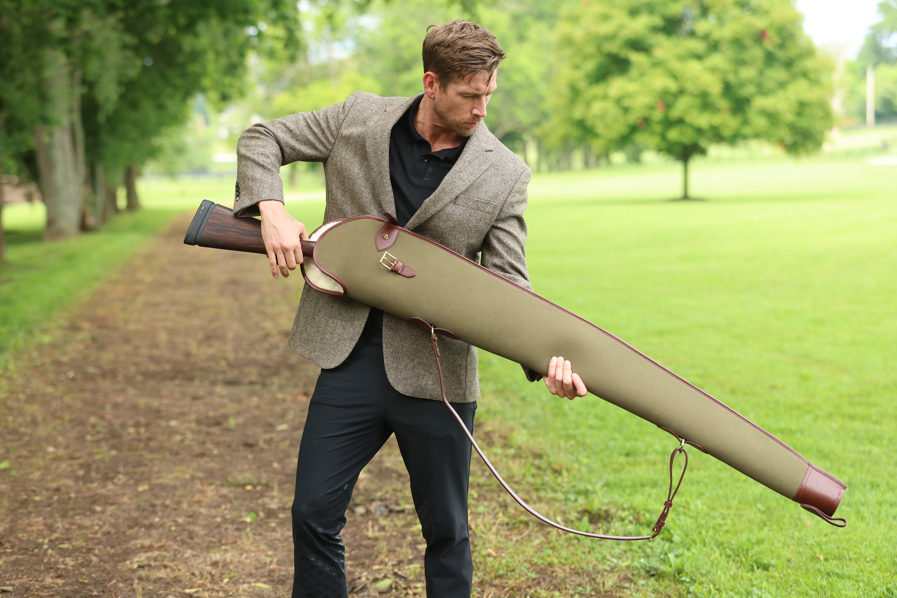 Man in a brown tweed jacket holding a Military Green Sandringham Gun Slip with Havana brown leather trim outdoors in a grassy park, made by Freedman’s Harness.