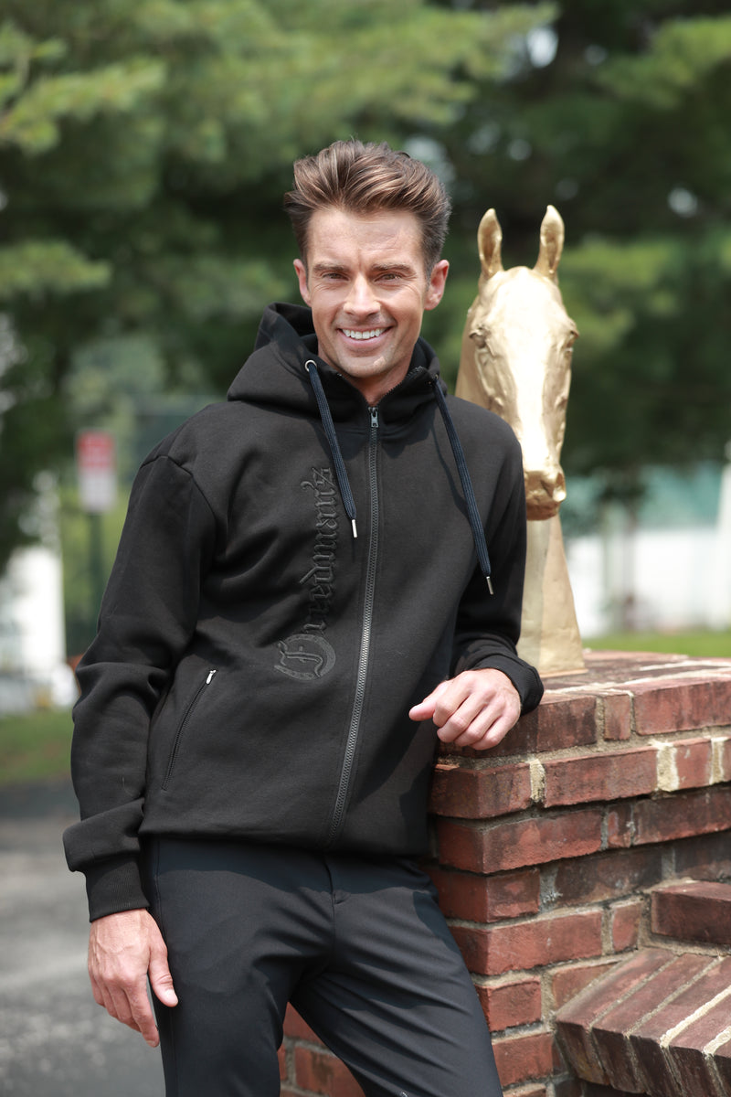 Smiling man wearing the Men's Black Logo Hoodie by Freedman's, standing outdoors next to a golden horse statue and a brick wall."