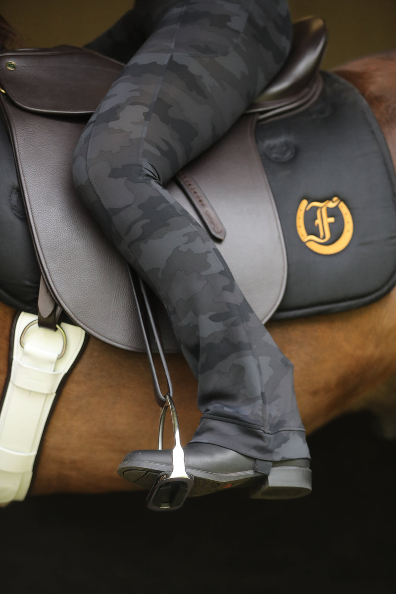 Tous les Jours Performance Jods, black camouflage pattern, worn by rider seated on a horse with dark leather saddle and silver stirrups, circular logo on saddle pad, outdoor setting.