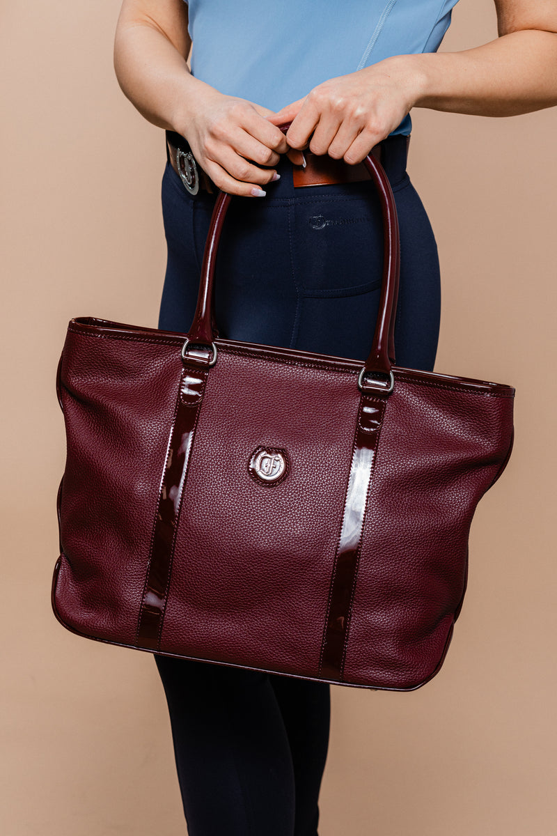 Equestrian-inspired elegance: The Victoria Tote by Freedman's in Burgundy, a luxurious leather handbag with glossy patent accents, carried by a stylish rider dressed in navy breeches and a light blue top, against a neutral beige background