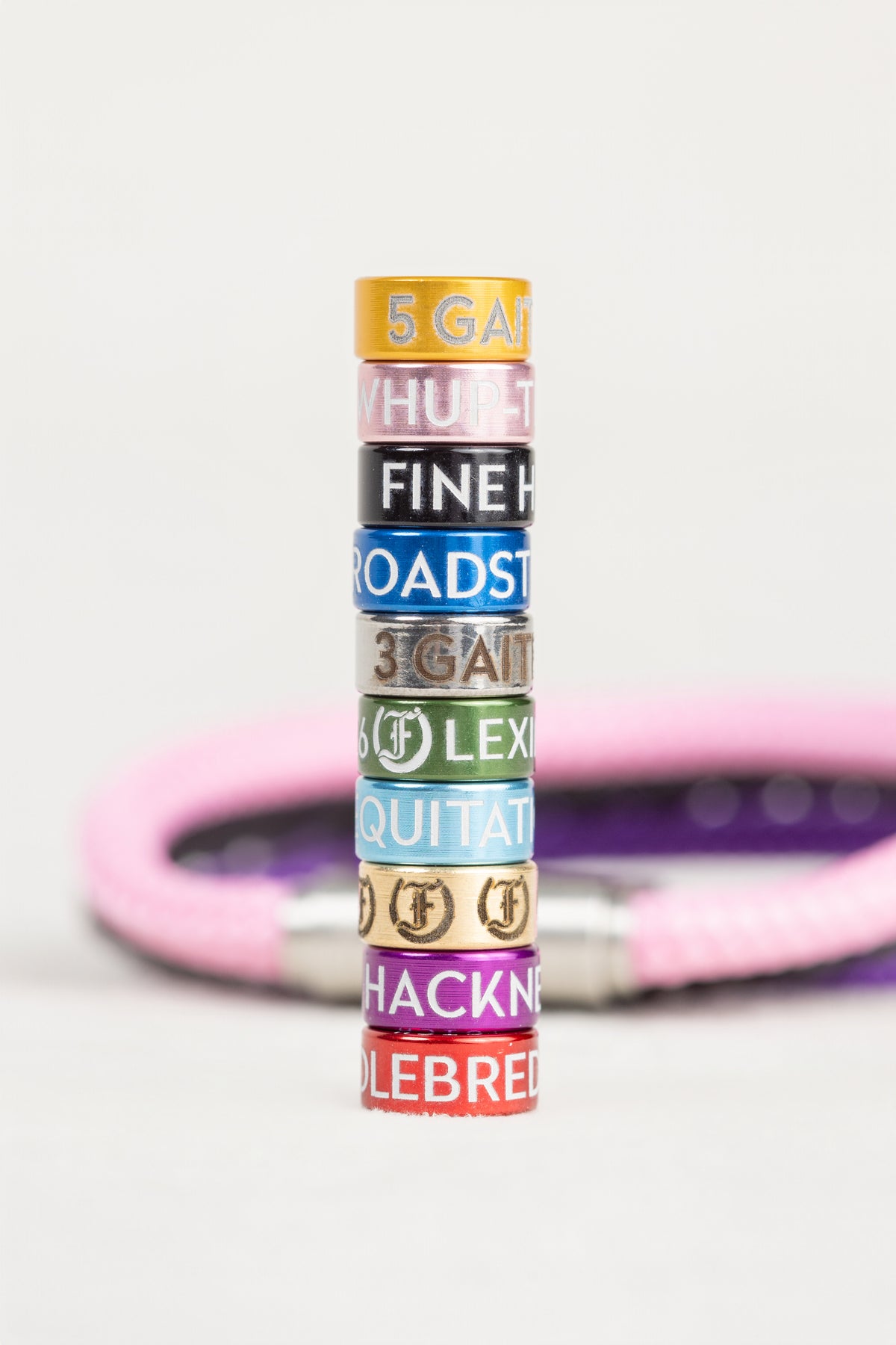 A vertical stack of nine colorful leather bangles with engraved horse racing names and numbers, set against a blurred pink leather bracelet on a light gray background.