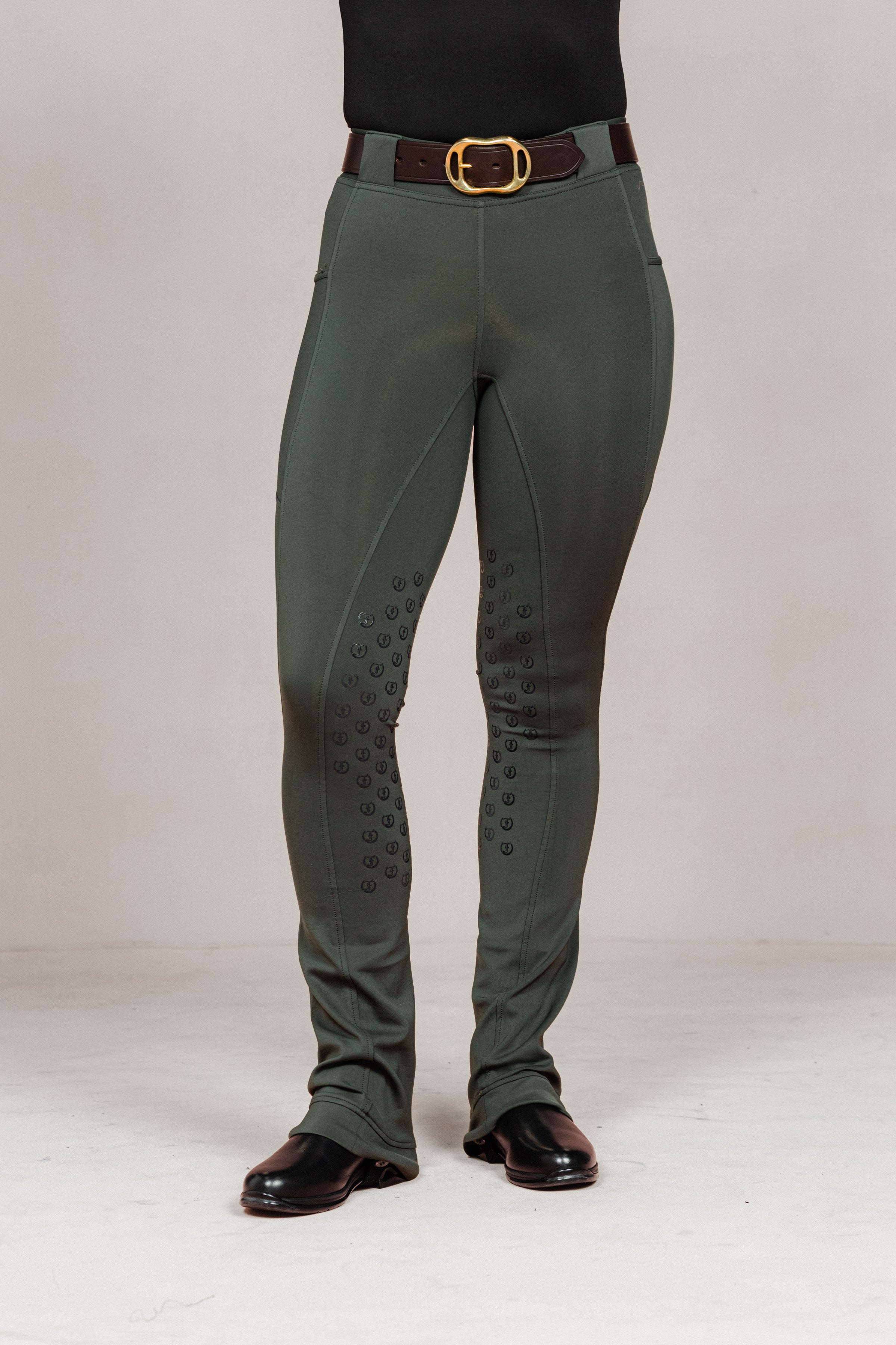 Front view of women's Umber Green jodhpurs by Freedman Harness, featuring full seat silicone grip, flared leg, brown belt with gold buckle, and black riding boots – high-performance equestrian pant