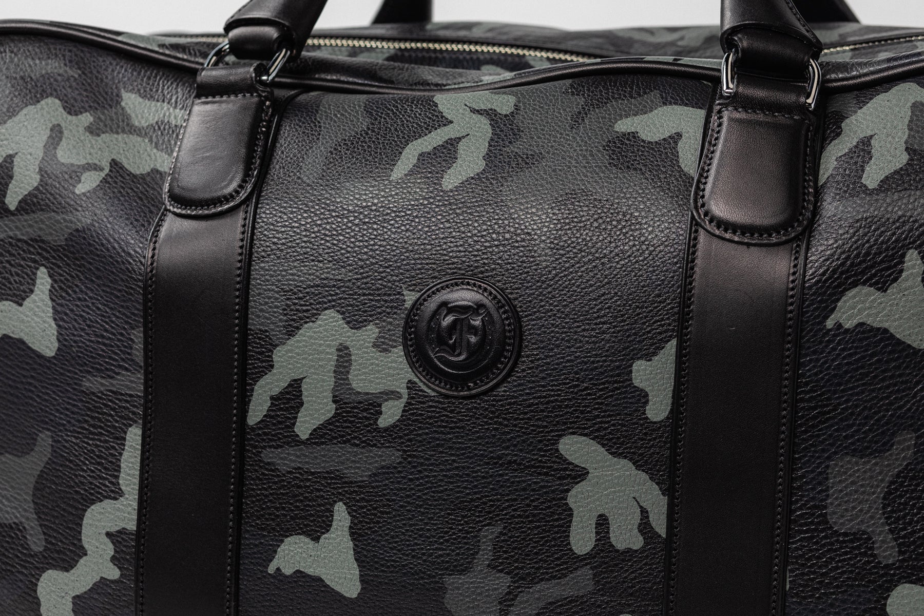 Close-up of Freedman's Grand Break Duffel, center detail, embossed "F" logo on dark leather panel, camouflage patterned leather with visible texture, dark leather trim along sides, silver hardware visible, light background