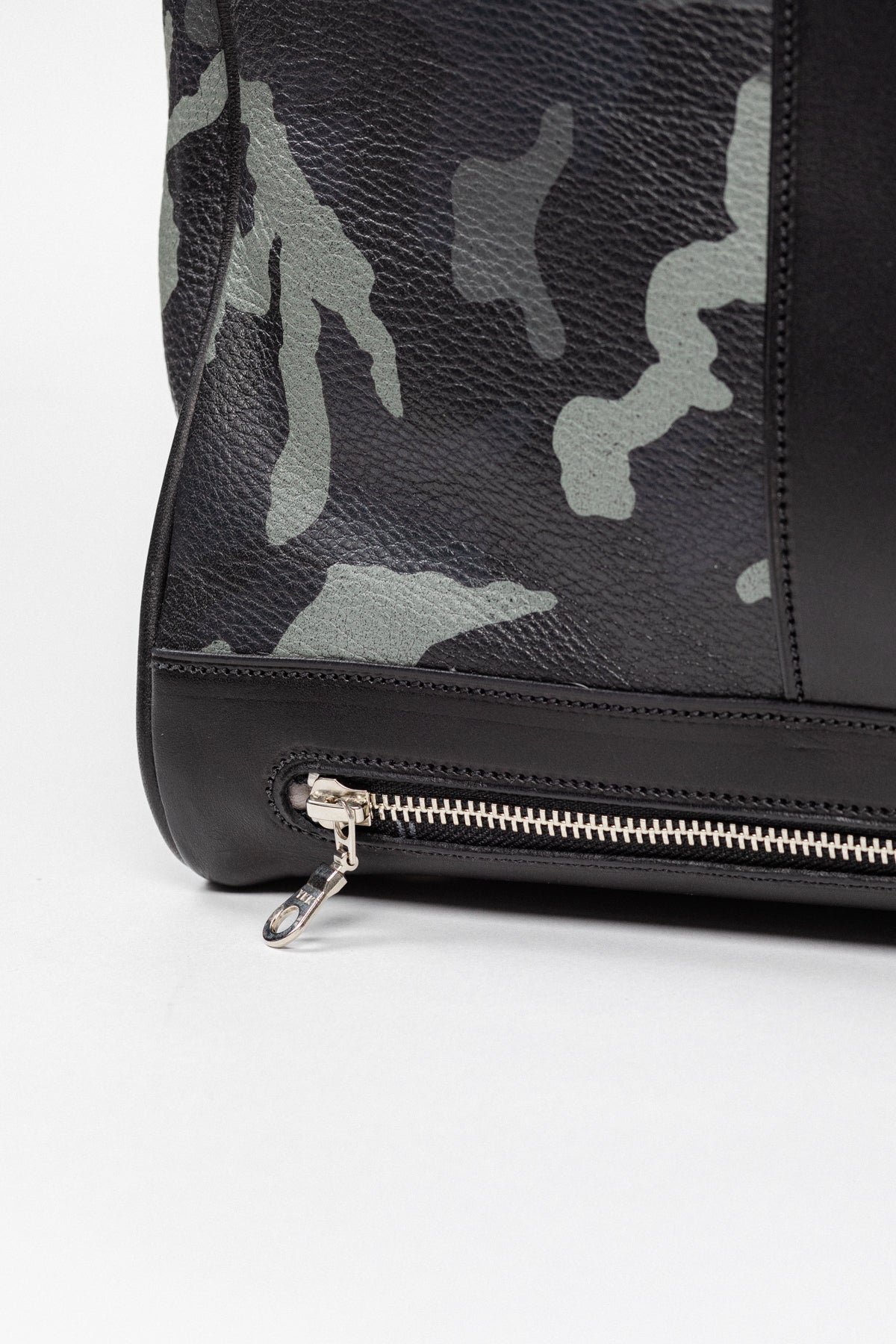 Close-up of Freedman's Grand Break Duffel, bottom corner detail, camouflage patterned leather, dark leather trim along base, silver zipper with elongated pull, light background.