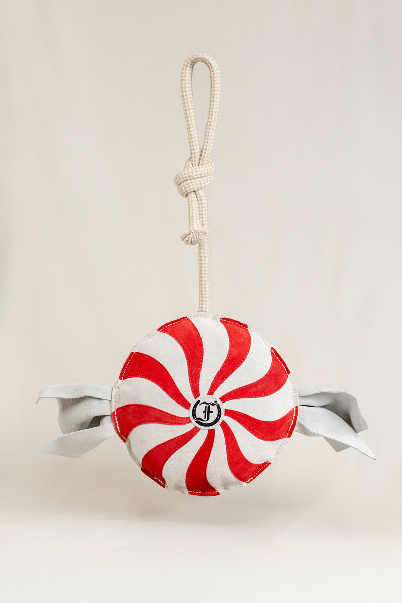 Red-and-white peppermint swirl horse toy with fabric ends and a rope handle, featuring the Freedman logo at the center, displayed against a beige background."