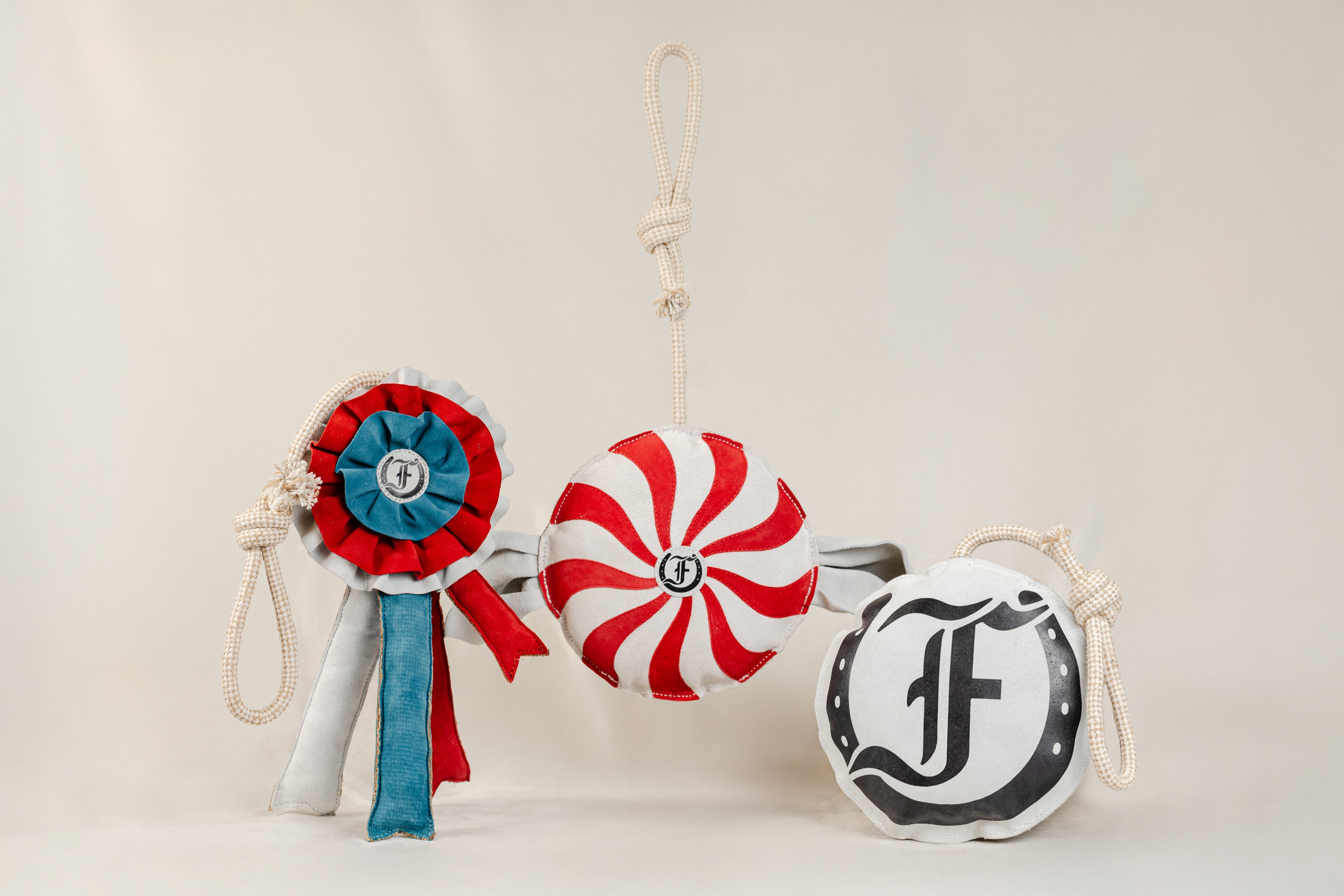Collection of handmade equestrian horse toys including a red, blue, and white ribbon rosette, a peppermint-style red-and-white ball, and a black-and-white Freedman logo toy, each with a rope handle, shown on a beige background