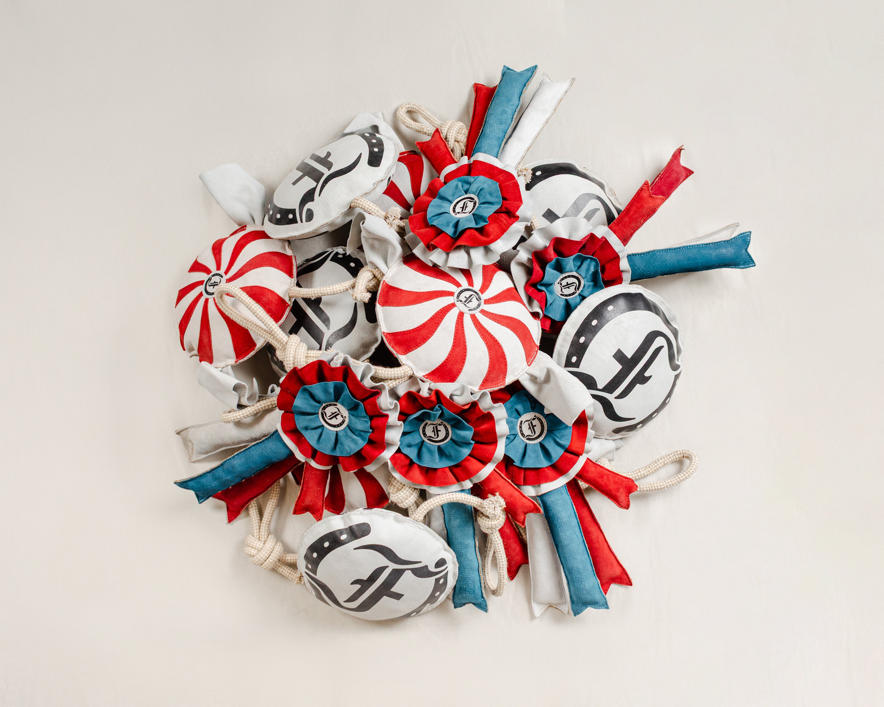 Pile of handcrafted equestrian horse toys featuring red, blue, and white ribbon rosettes, red-and-white peppermint balls, and black-and-white Freedman logo toys, all with rope handles, displayed on a beige background