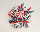Pile of handcrafted equestrian horse toys featuring red, blue, and white ribbon rosettes, red-and-white peppermint balls, and black-and-white Freedman logo toys, all with rope handles, displayed on a beige background