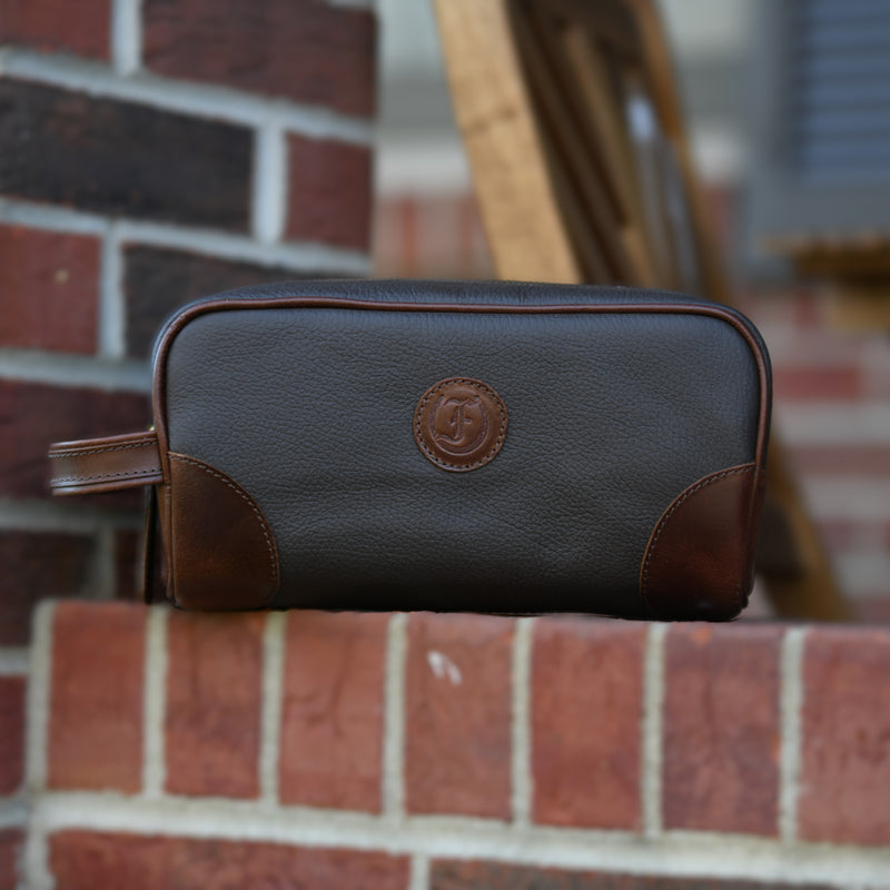 Freedman's Hansom Shave Bag, dark brown and tan two-tone leather, embossed "F" logo on front, side handle, outdoor setting with brick and wood background.