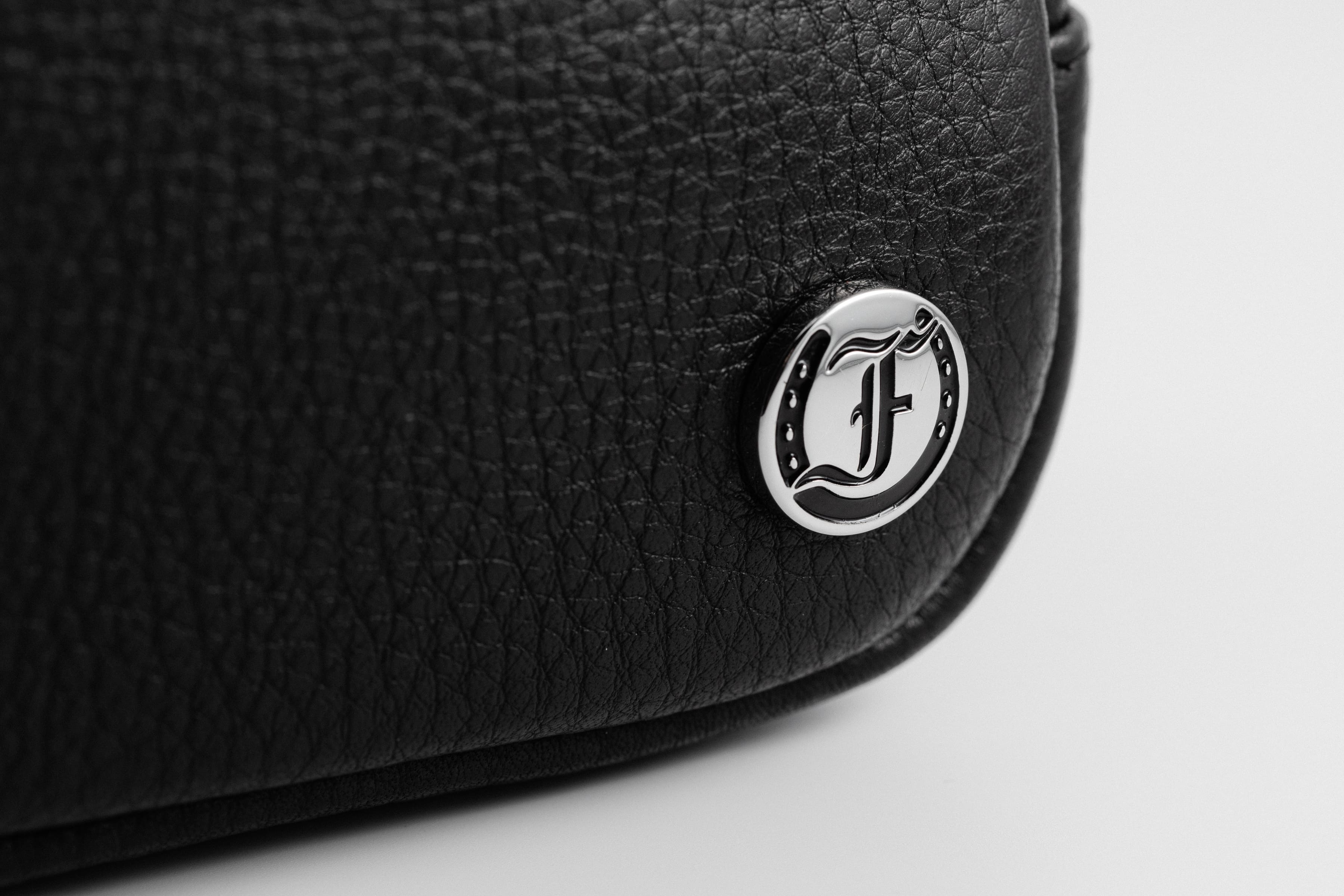 Close-up of Freedman's Norfolk Crossbody Bag in Black Leather, showing silver logo medallion on textured leather.