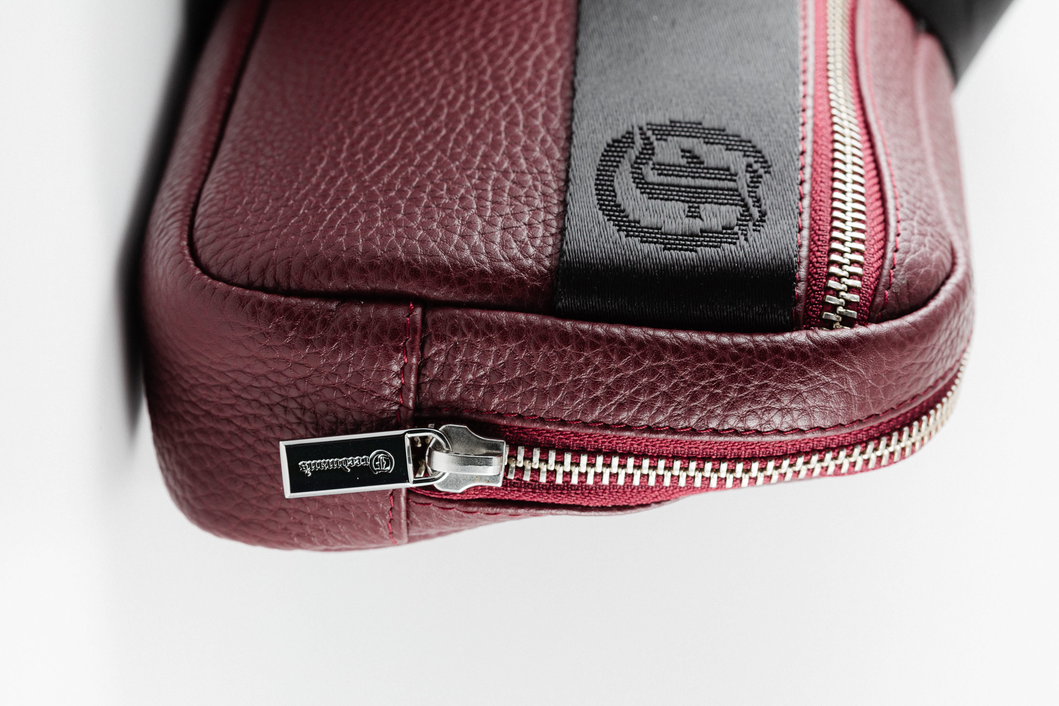 Close-up of Freedman's Norfolk Crossbody Bag in Burgundy Leather, showing silver zipper, logo pull, and branded fabric strap.