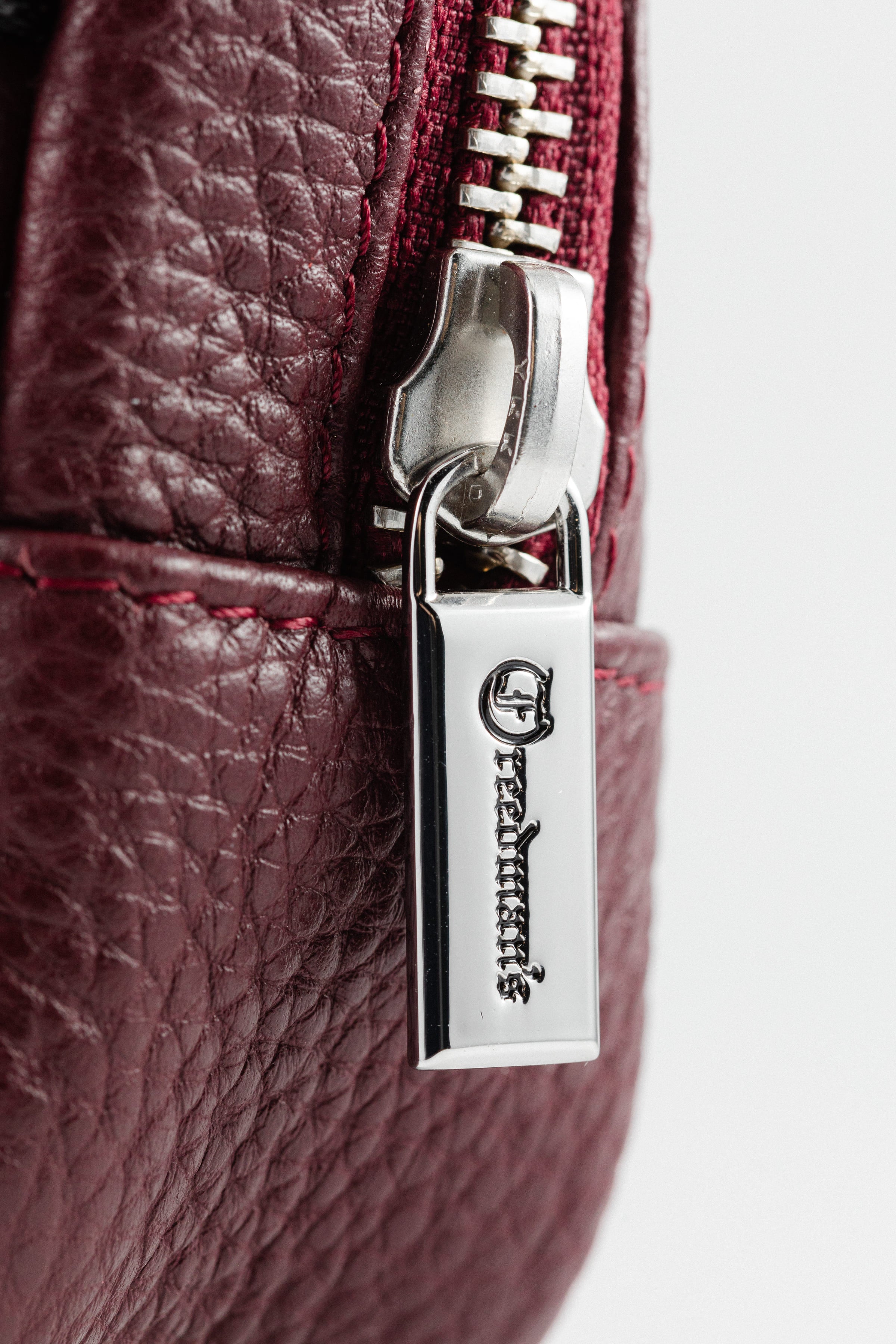 Close-up of Freedman's Norfolk Crossbody Bag in Burgundy Leather, showing silver zipper, logo pull, and branded fabric strap.