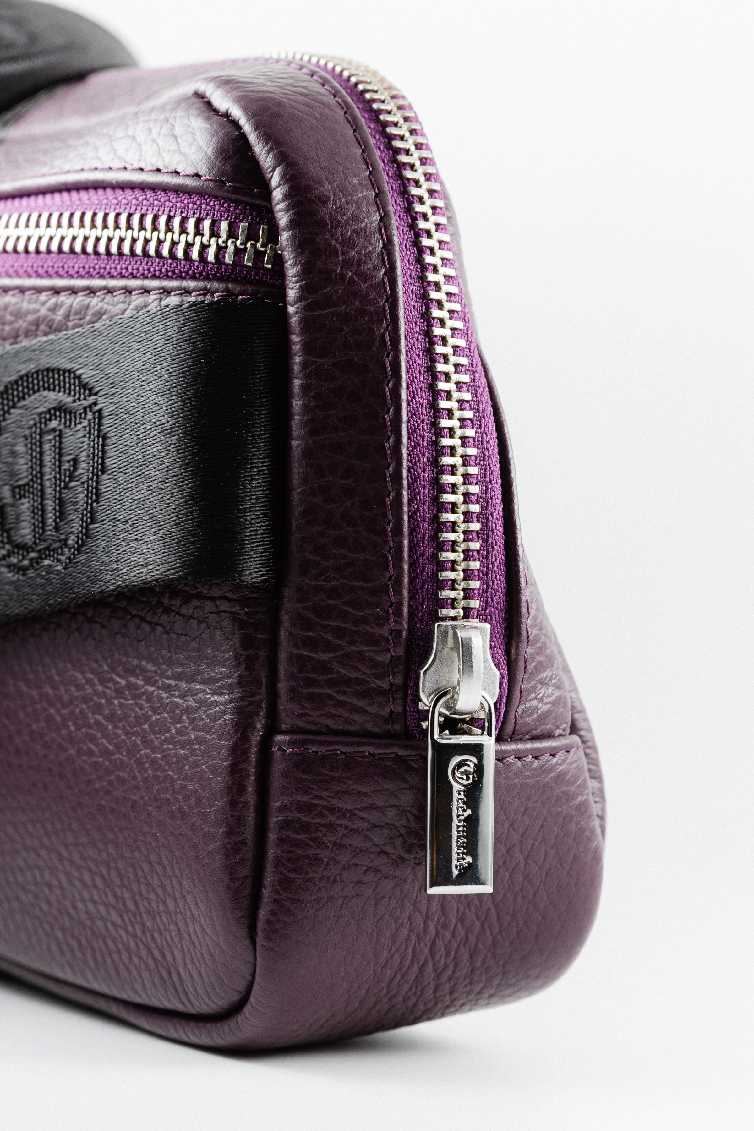 Close-up of Freedman's Norfolk Crossbody Bag in Purple Leather, showing silver zipper, logo pull, and branded fabric strap.