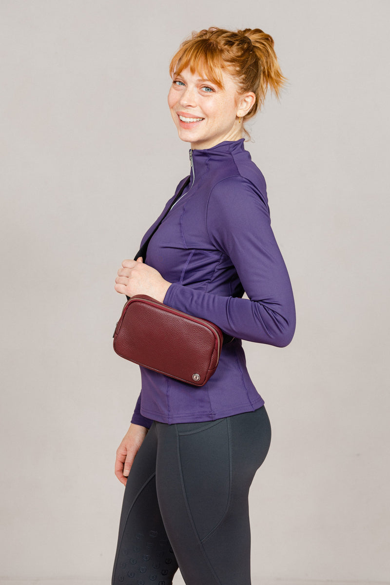 A smiling woman wearing the Norfolk Crossbody Bag in Black Leather by Freedman’s. She is dressed in a fitted purple athletic zip-up top and dark Jods.