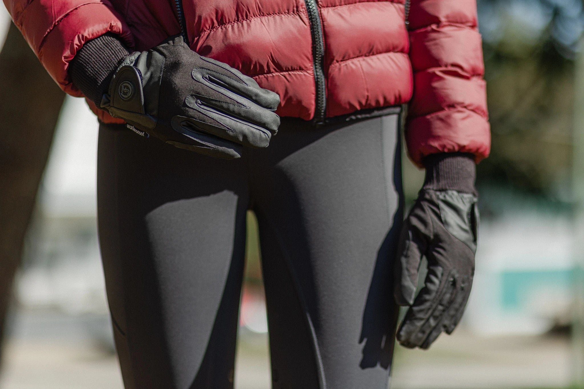 Close-up of Signature Ladies' II Winter Gloves in black, showing the reinforced fingers, adjustable wrist straps, and ribbed knit cuffs with a burgundy puffer jacket.