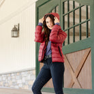 Woman wearing the Signature Ladies' Packable Puffer Jacket in burgundy with a hood, standing in front of a stable.