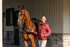 Woman wearing the Signature Ladies' Packable Puffer Jacket in burgundy while holding a saddled horse outside a stable.