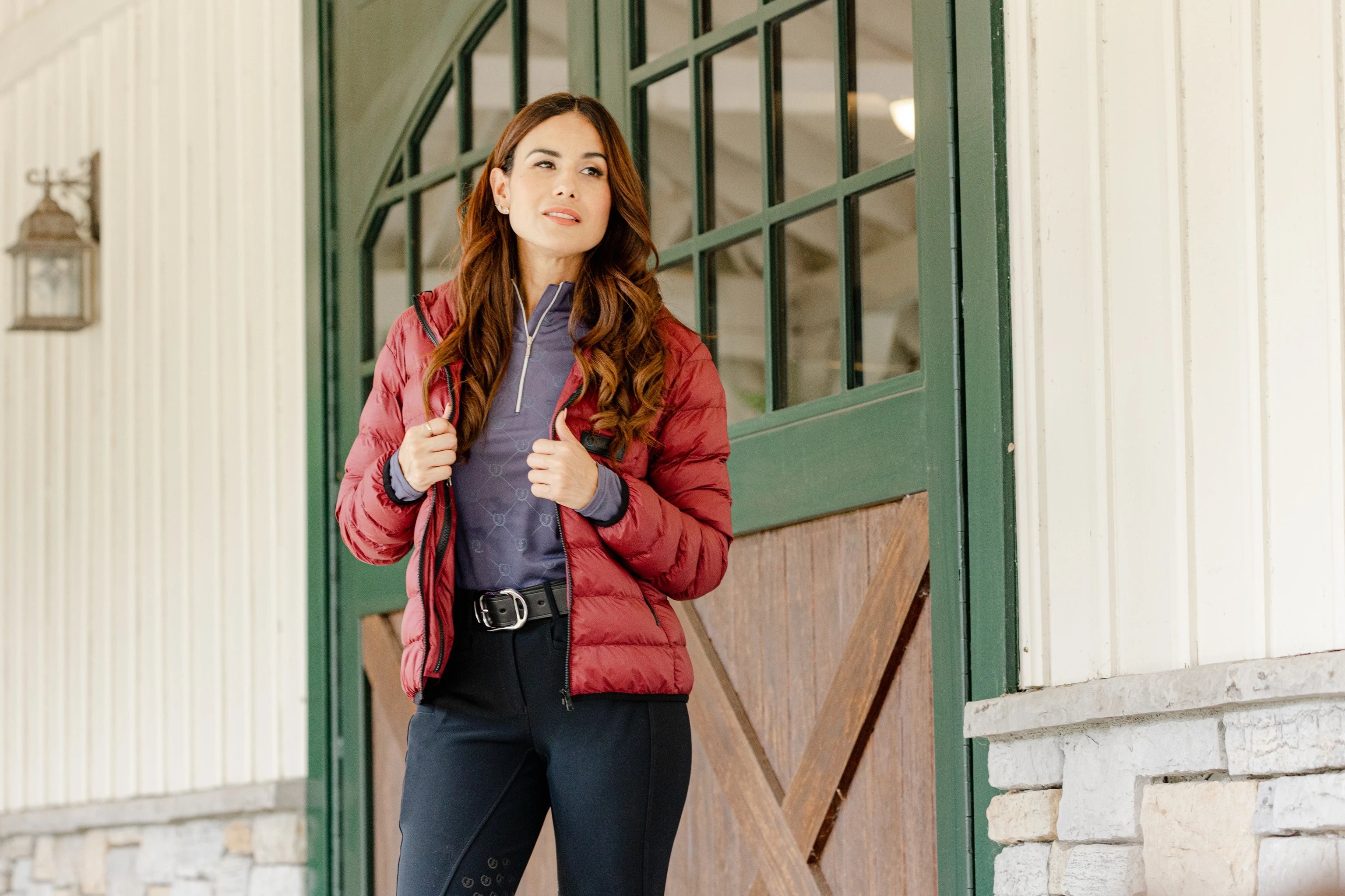 Woman wearing the Signature Ladies' Packable Puffer Jacket in burgundy, featuring a quilted design, front zipper, zippered pockets, and hood, leaning against a stable door.