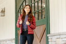 Woman wearing the Signature Ladies' Packable Puffer Jacket in burgundy, featuring a quilted design, front zipper, zippered pockets, and hood, leaning against a stable door.