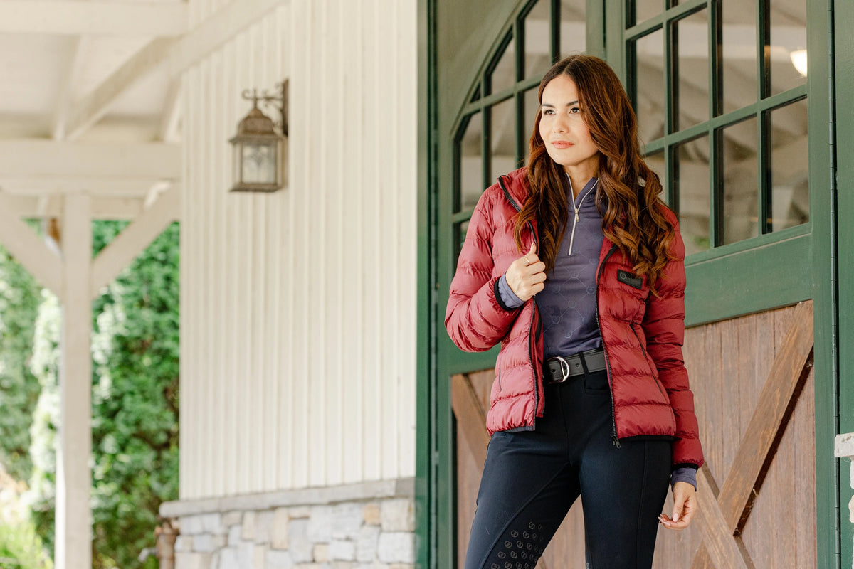 Woman wearing the Signature Ladies' Packable Puffer Jacket in burgundy, featuring a quilted design, front zipper, zippered pockets, and hood, leaning against a stable door.